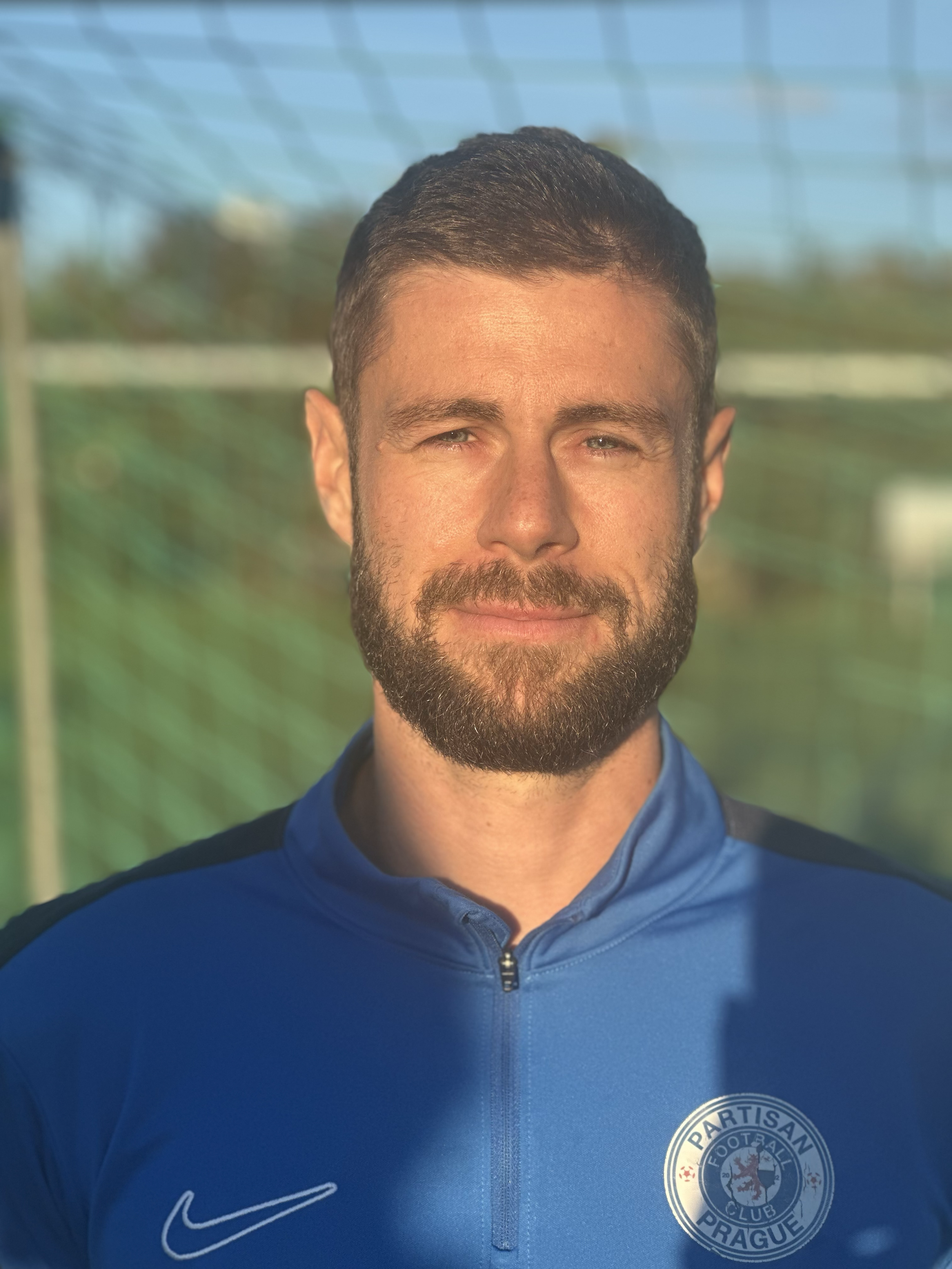 A man with a beard and short hair wearing a blue sports jacket with a Partisan Prague football club logo, standing outdoors in sunlight with a blurred background.