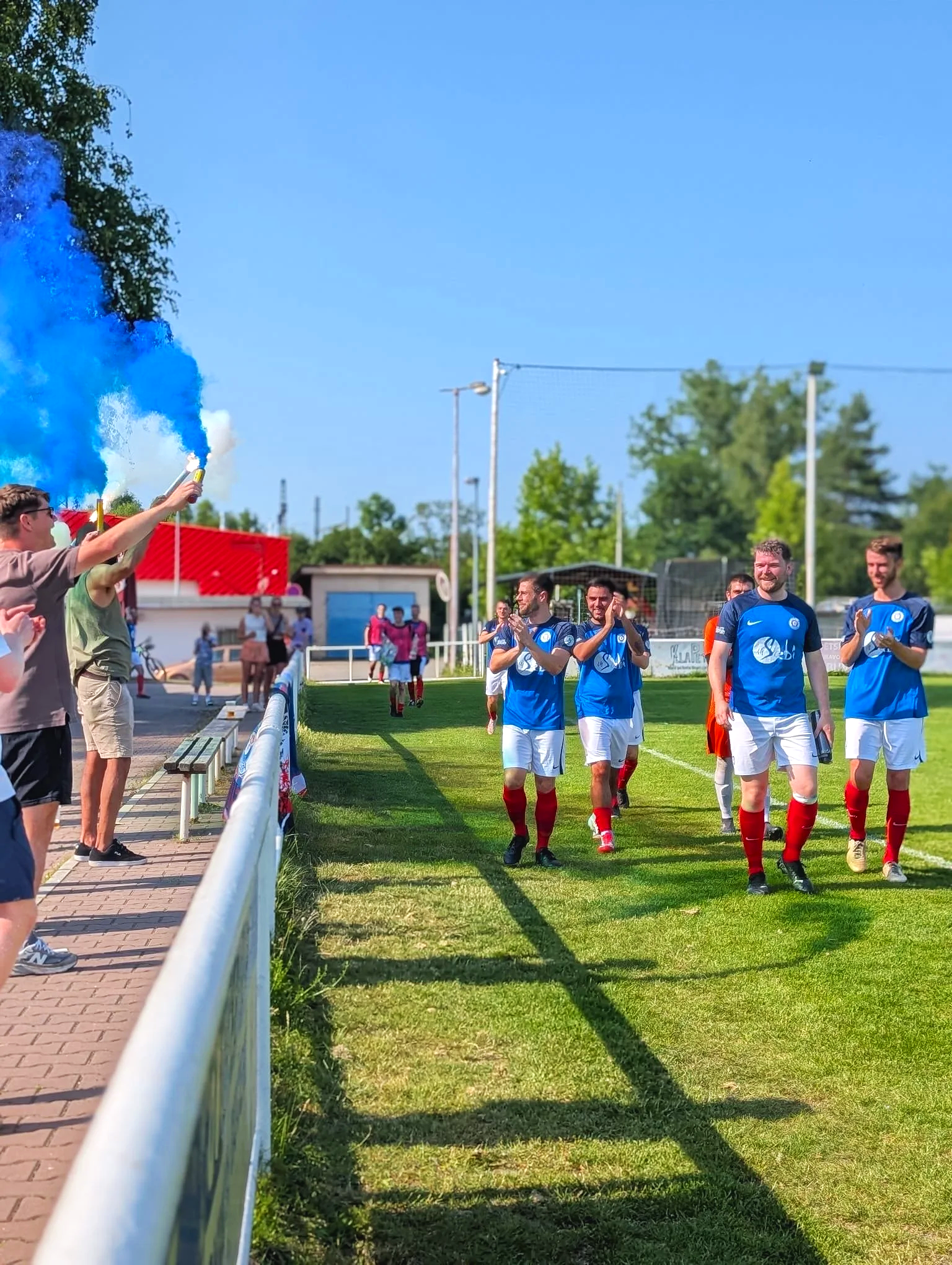Soccer players in blue jerseys and white shorts walking onto the field while fans cheer and hold blue smoke flares.