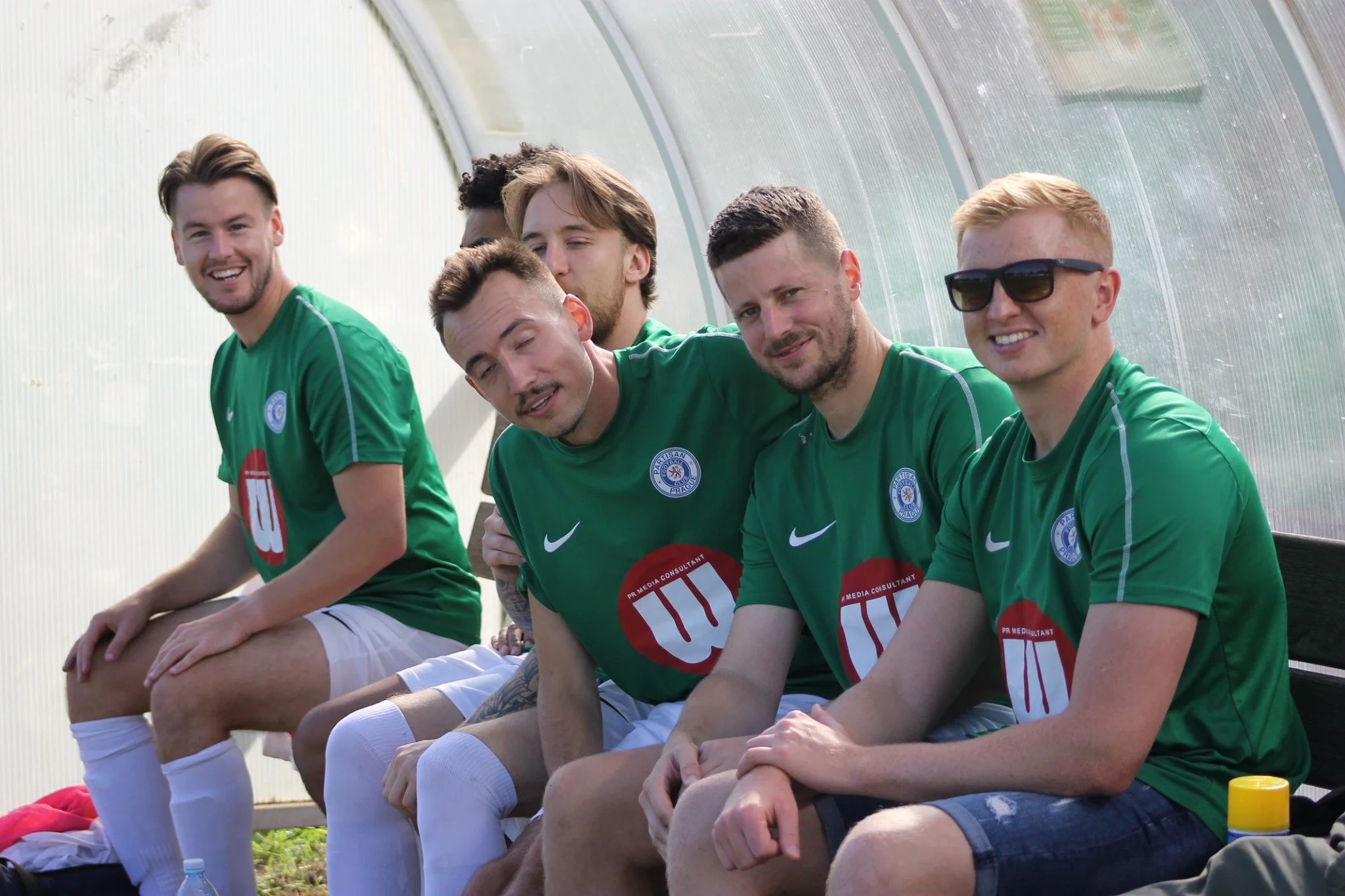 Six young men sitting on a bench in green soccer jerseys, inside a sports team's dugout or bench area, smiling and posing for the photo.