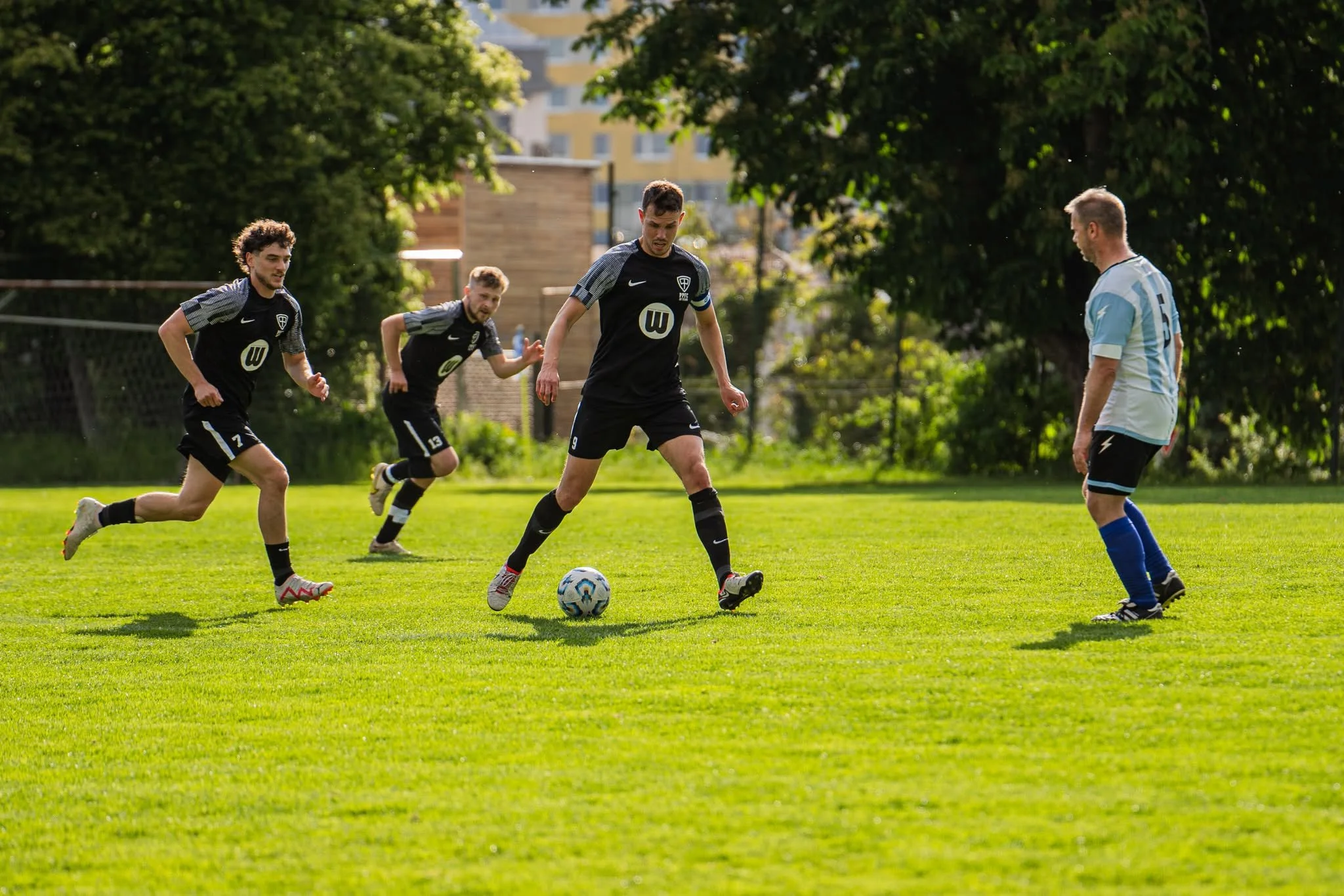 Four men playing soccer on a grassy field during daytime. One man in a light blue and white striped jersey faces a man in a black jersey with the number 9. Two other men in black jerseys are running in the background, chasing the ball.