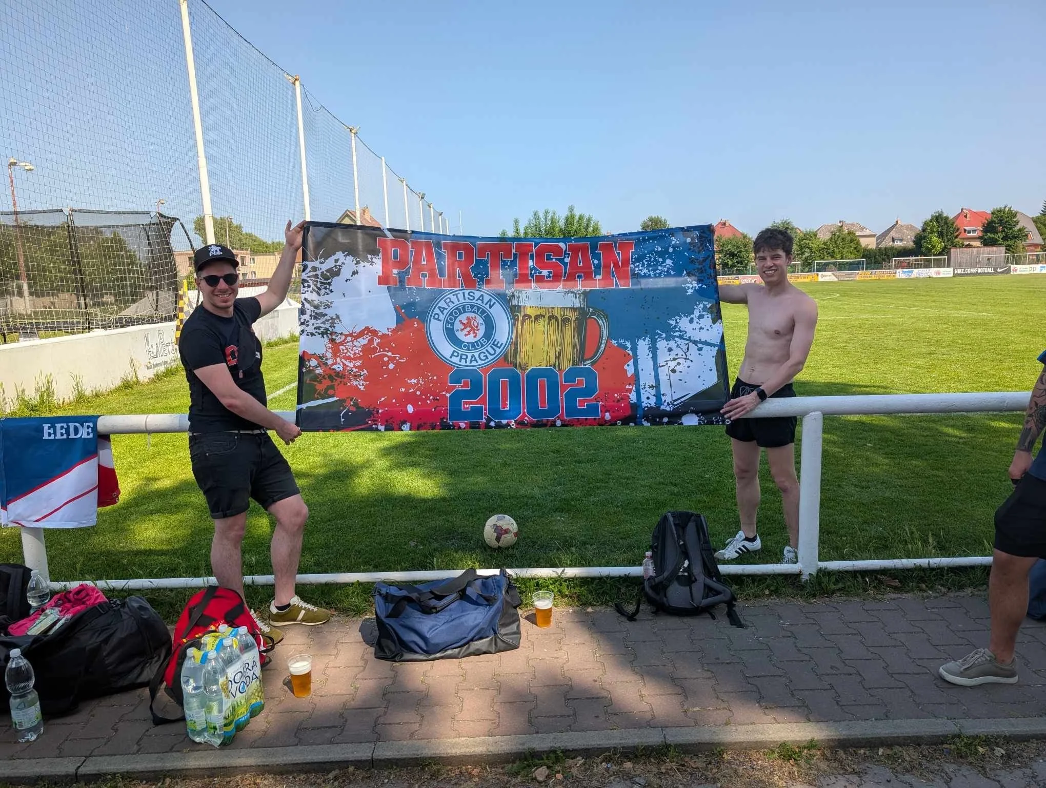 Two young men stand on a soccer field, holding a banner that reads 'PARTISAN 2002' with a logo and a beer mug illustration, celebrating a soccer event in 2002. One man is shirtless, and the other wears a black t-shirt, shorts, and sunglasses. Soccer
