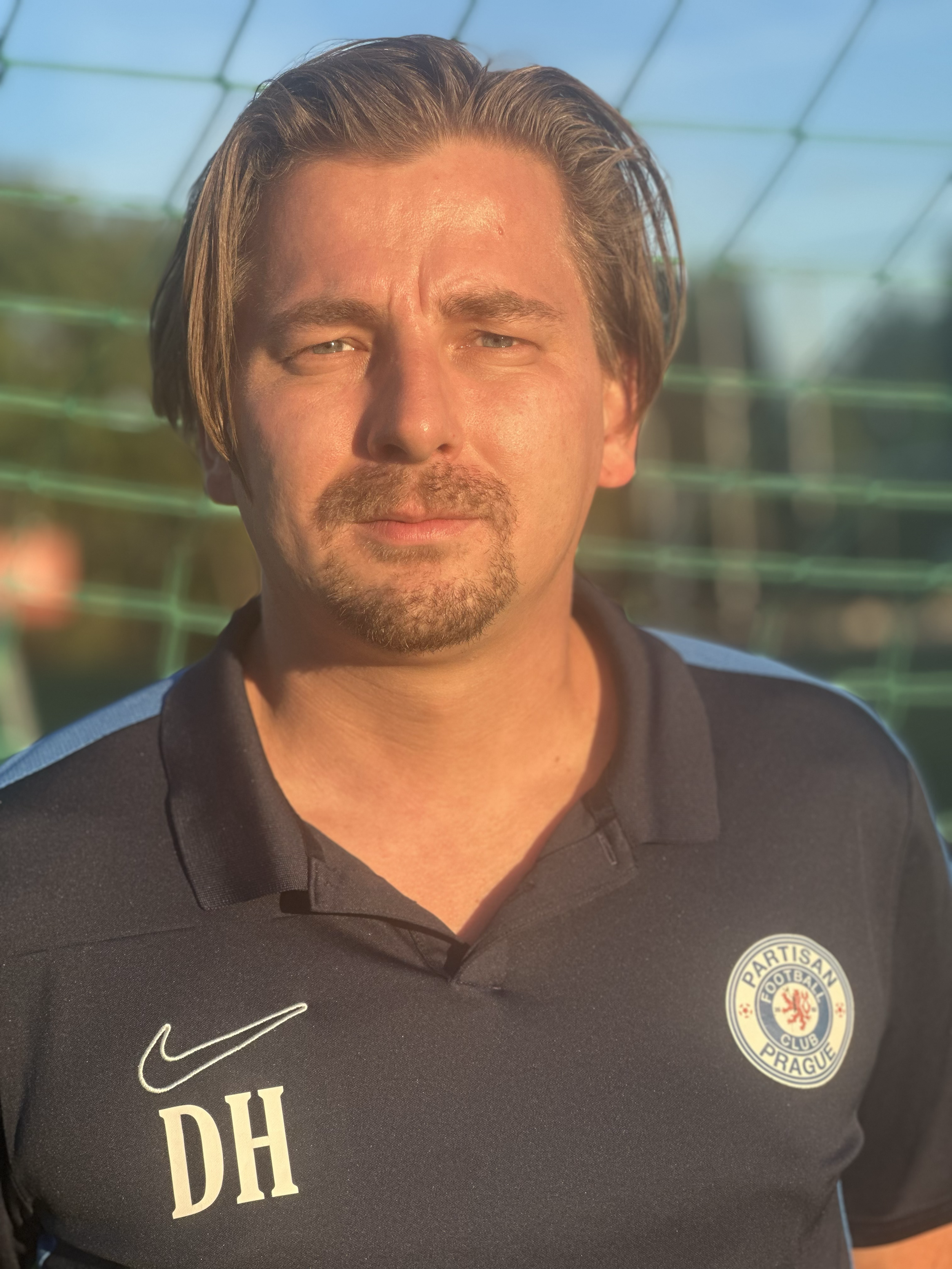 A man with brown hair and a goatee looks into the camera, wearing a black sports polo shirt with patches including a logo of the Prague Football Club and initials 'DH'. The background shows a sunny outdoor setting with a green net.