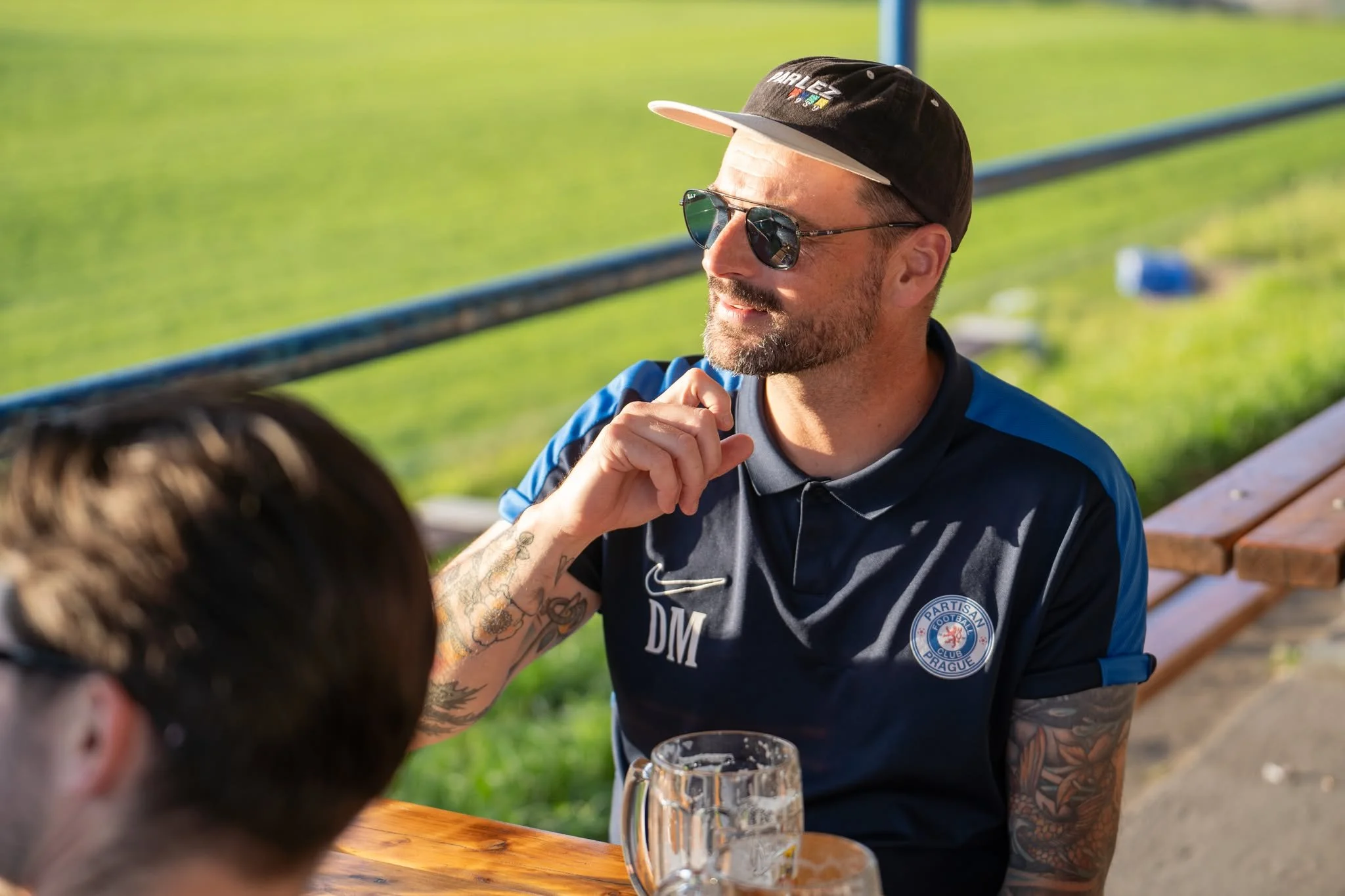 A man wearing sunglasses, a black cap, and a blue sports shirt with a logo, sitting at a wooden table outdoors with a green grassy background and a beer mug in front of him, engaged in conversation.