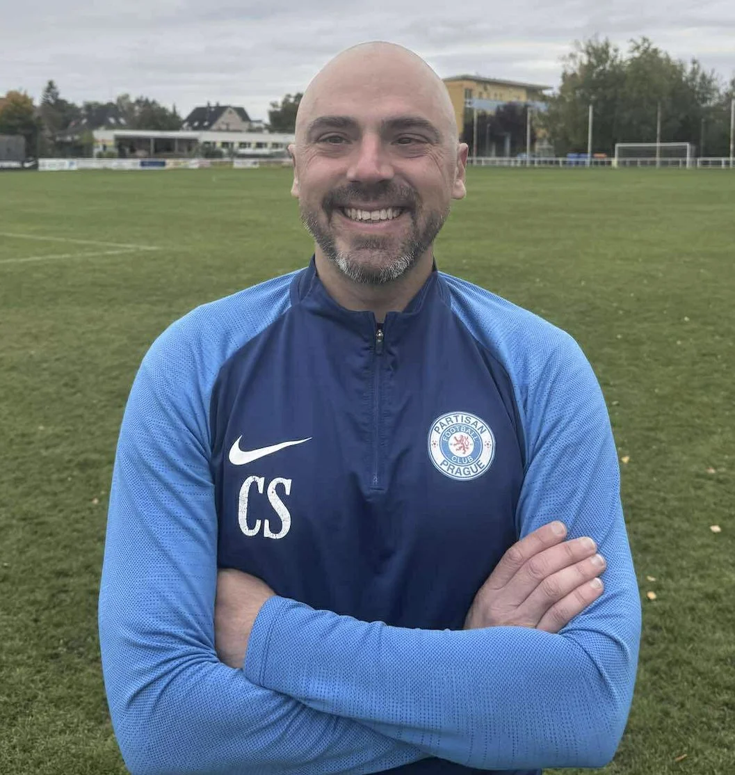 Male soccer player in white and blue uniform kicking a soccer ball during practice on a grass field.