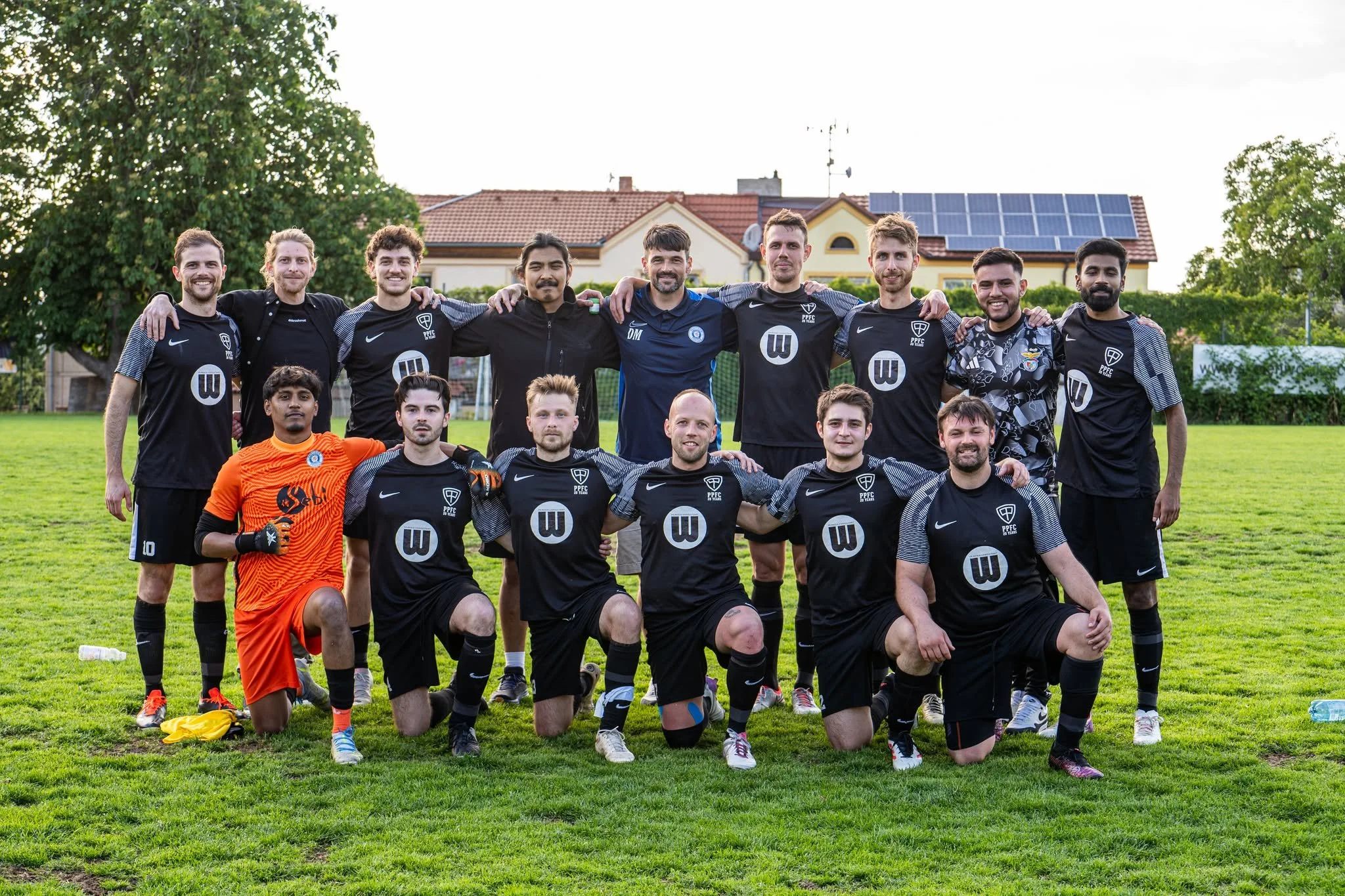 A group of 14 soccer players and coaches posing on a grassy field, with some kneeling in front and others standing behind, wearing black uniforms with a sponsor logo, and one player in an orange goalkeeper jersey.