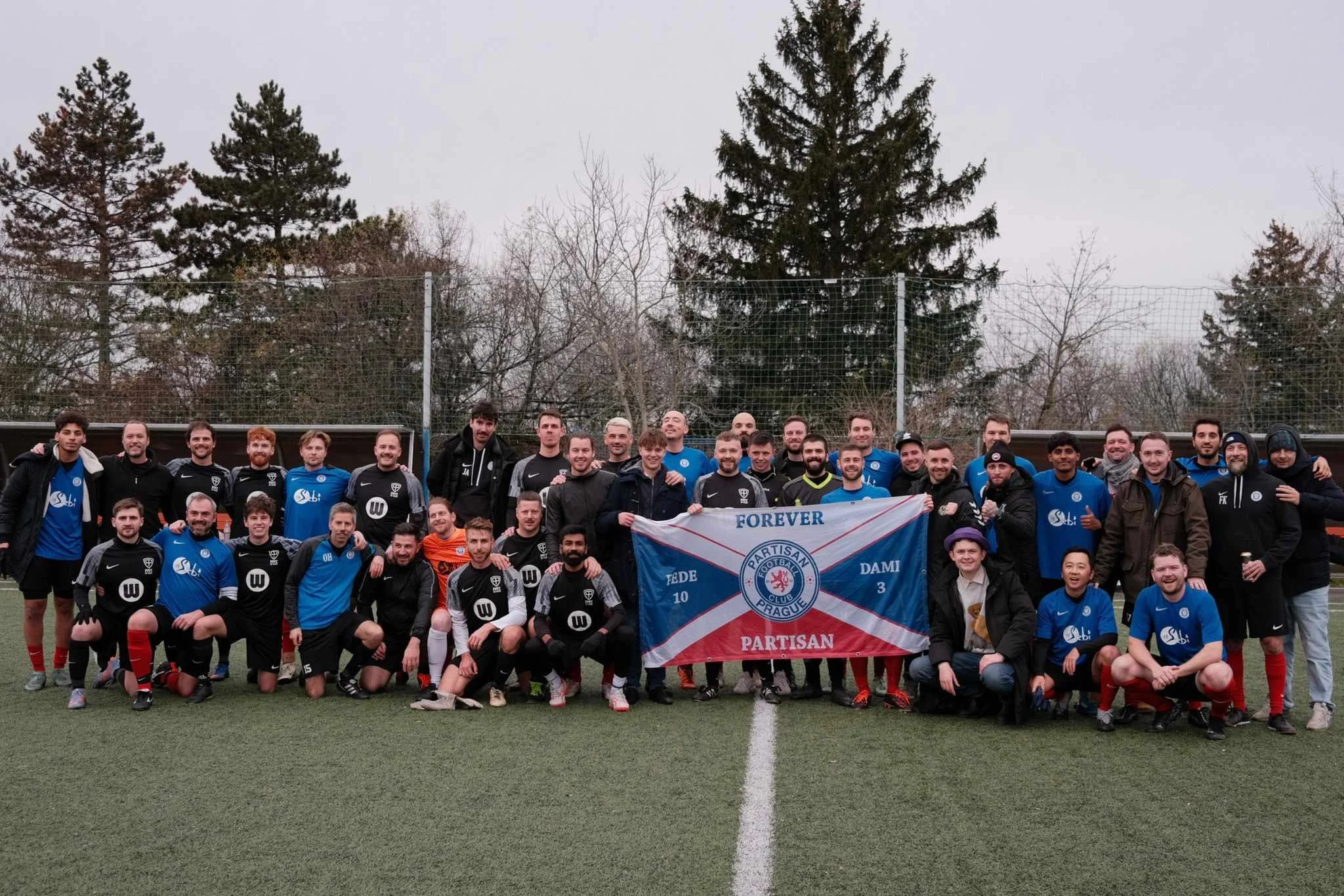Group of soccer players and supporters on a field holding a flag with a team logo, surrounded by trees, with gray sky overhead.