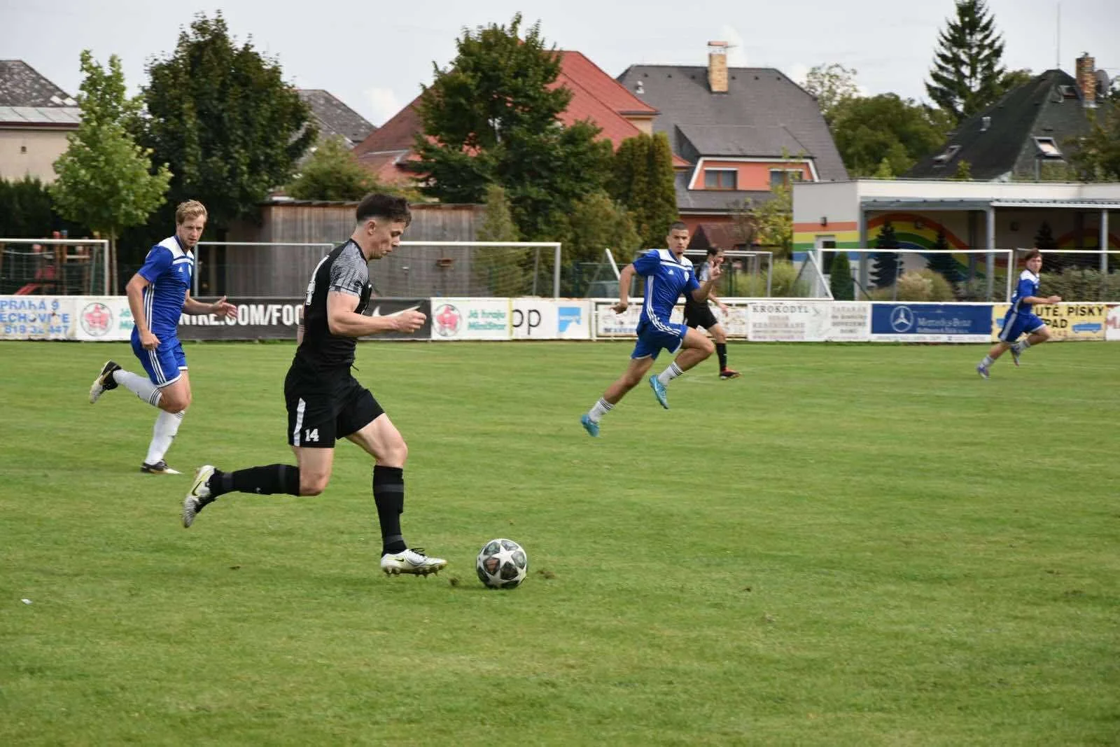Soccer players on a field during a match, with one player in black controlling the ball while others in blue chase behind.