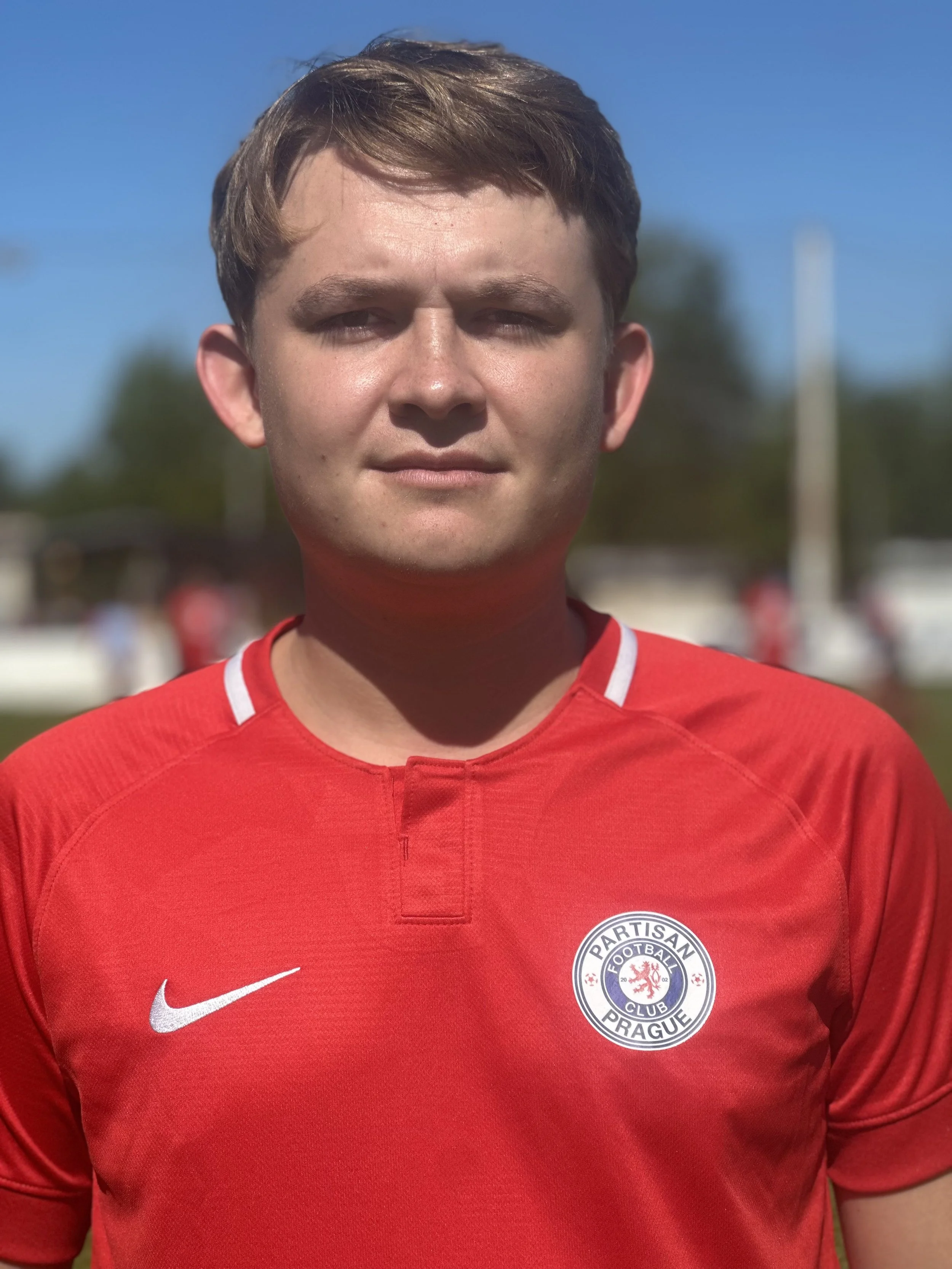 A young man wearing a red football jersey with the logo 'Partisan Football Club Prague' on the chest, standing outdoors under a clear blue sky.