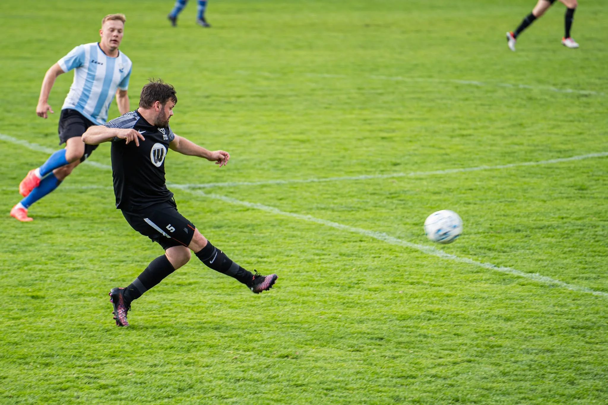 Two soccer players chasing after a ball on a green field, with one player in a black uniform and the other in a light blue and white striped jersey.