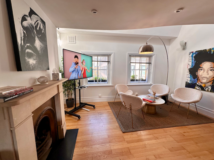 A bright and light apartment-style room with artwork on the walls and in front of two large windows, a soft area with table and white, fluffy chairs.
