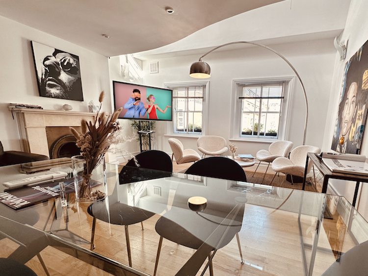 A bright and light apartment-style room with two separate areas, a large glass table in one half, surrounded by comfy chairs, while next to two large windows, a soft area with table and white, fluffy chairs.