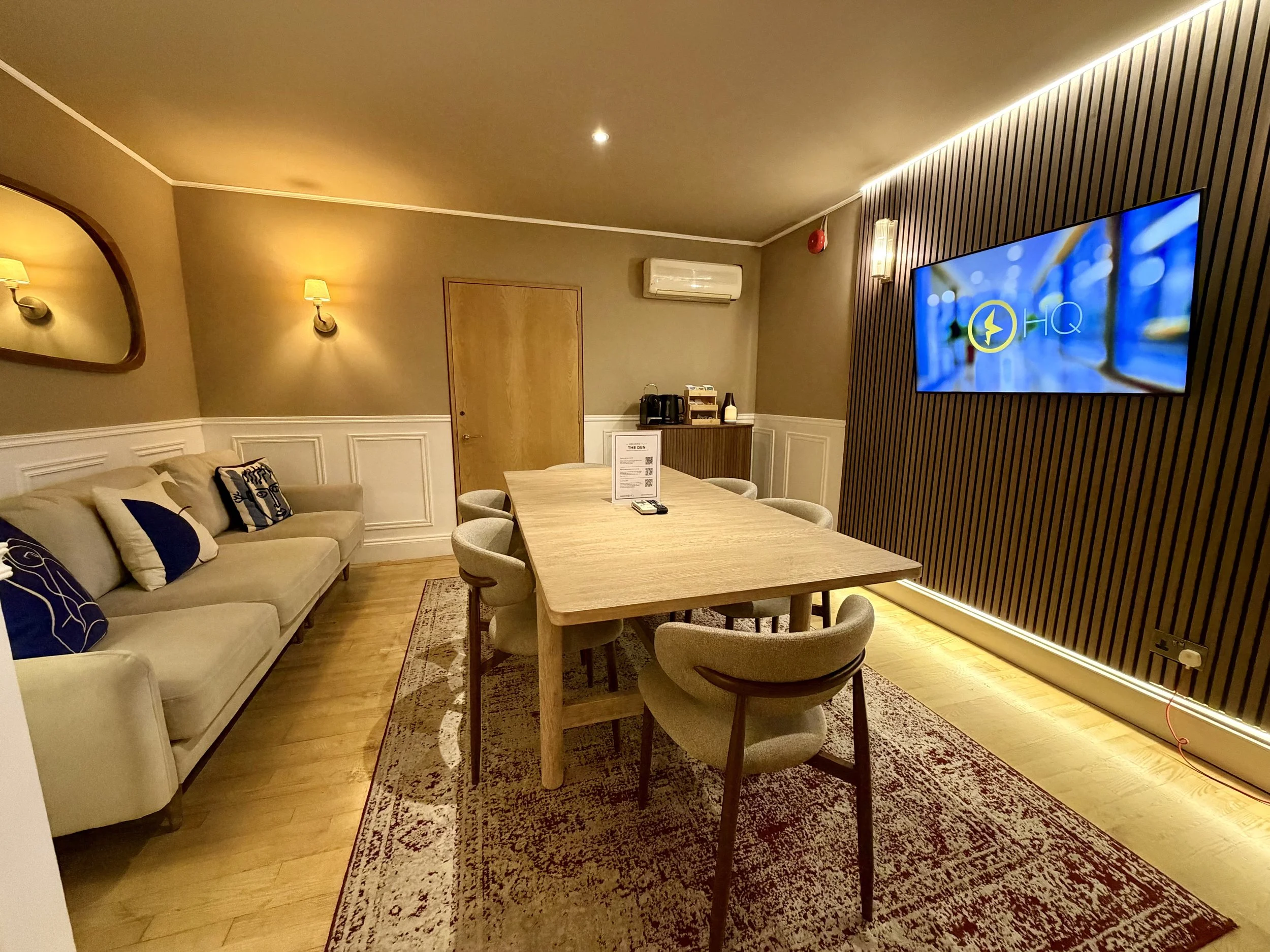 A boardroom with brown overtones, featuring a large oak table with chairs all around, a large TV attached to the wall and a generous sofa.