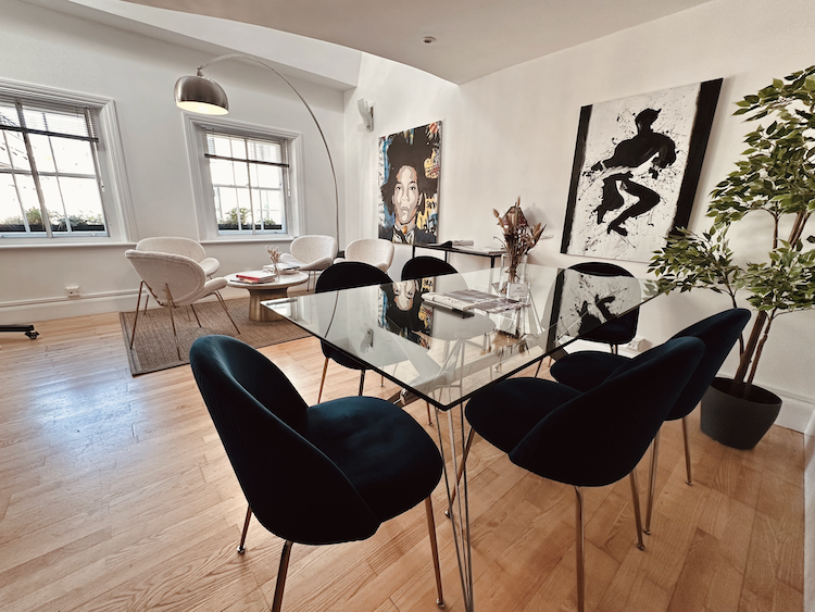 A bright and light apartment-style room with two separate areas, a large glass table in one half, surrounded by comfy chairs, while next to two large windows, a soft area with table and white, fluffy chairs.