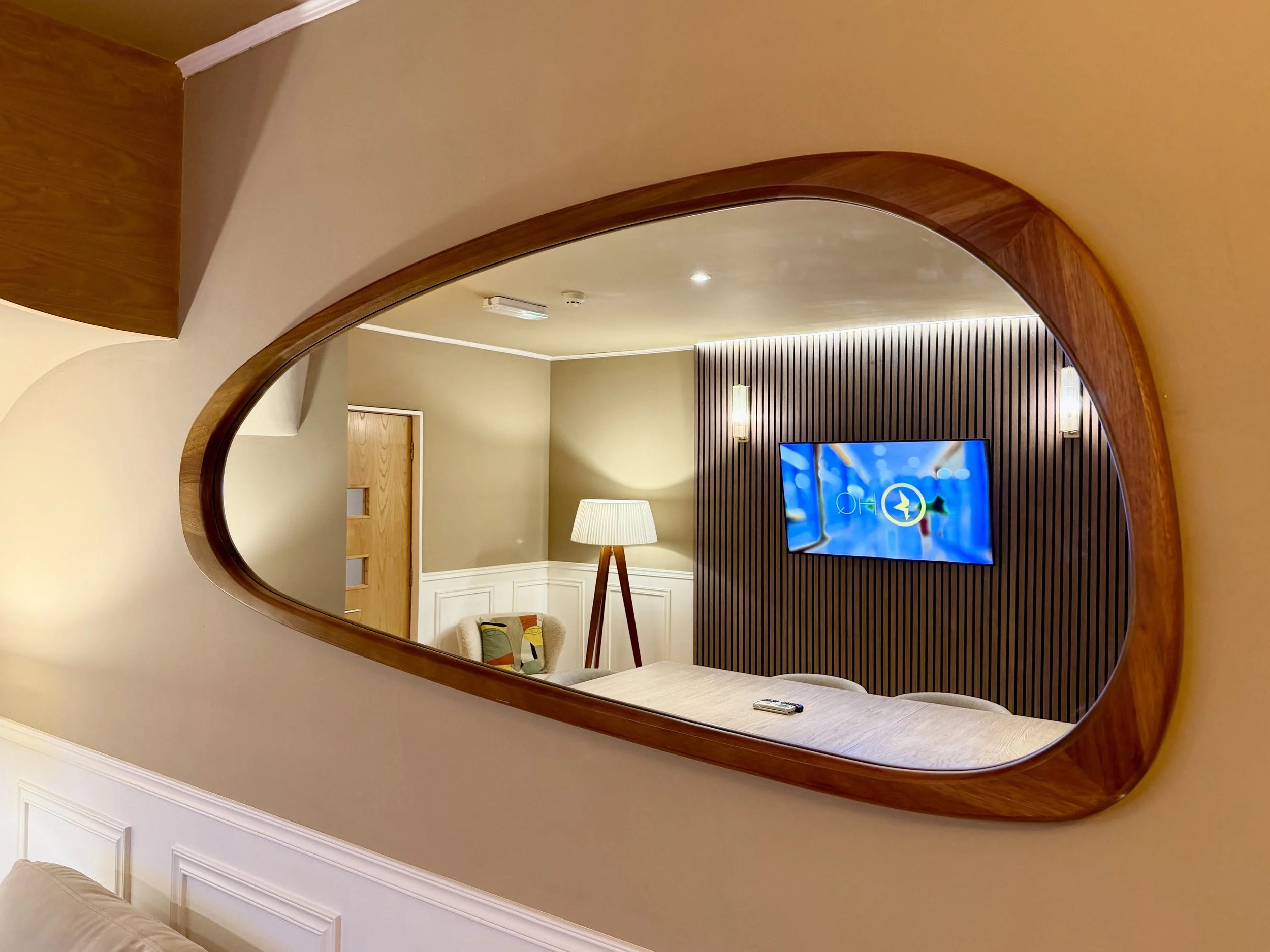 A mirror showing a boardroom with brown overtones, featuring a large oak table with chairs all around and a large TV attached to the wall in its reflection.