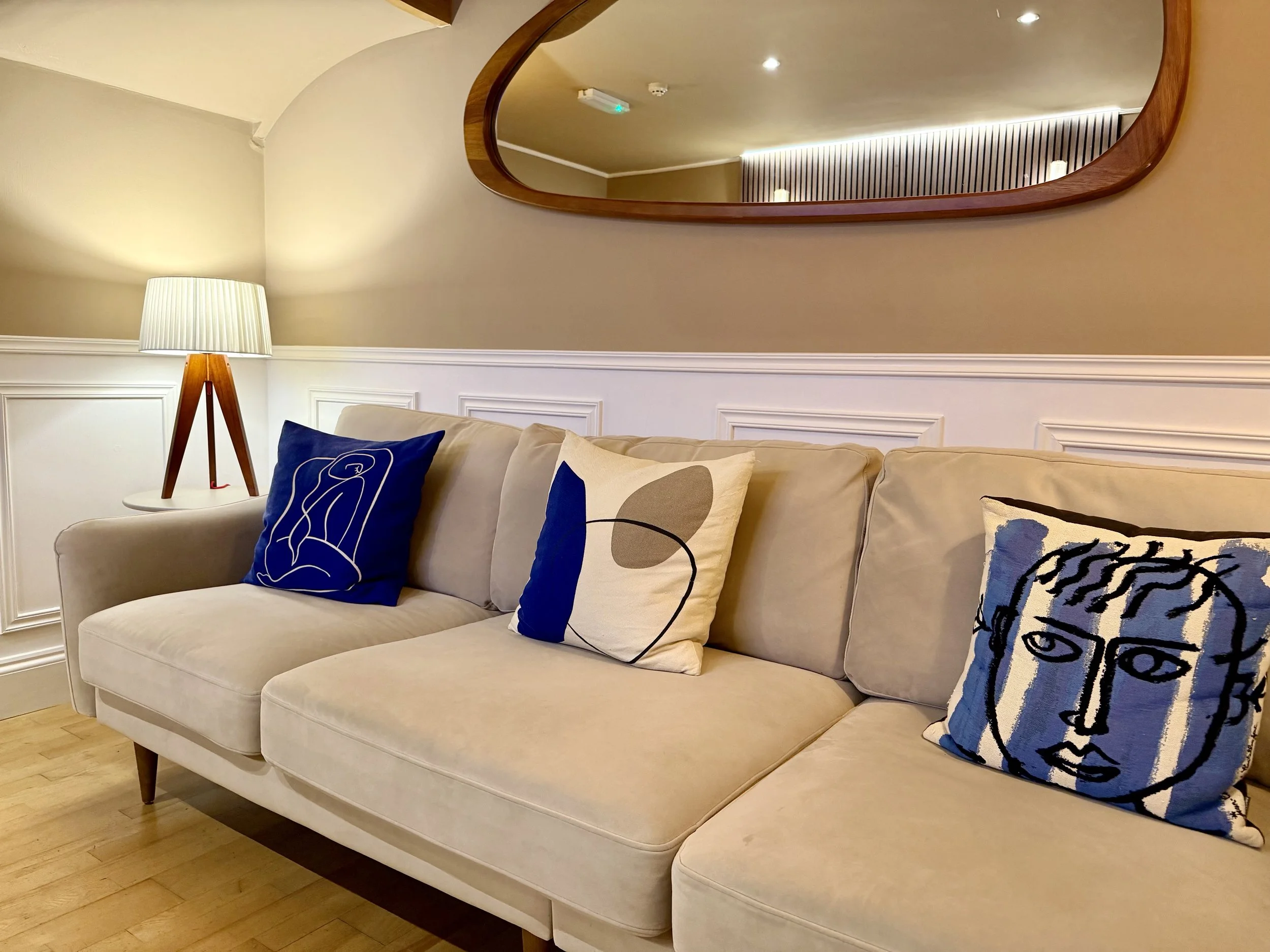 A big sofa with three cushions sits below a wall-mounted mirror and small lamp on a table.