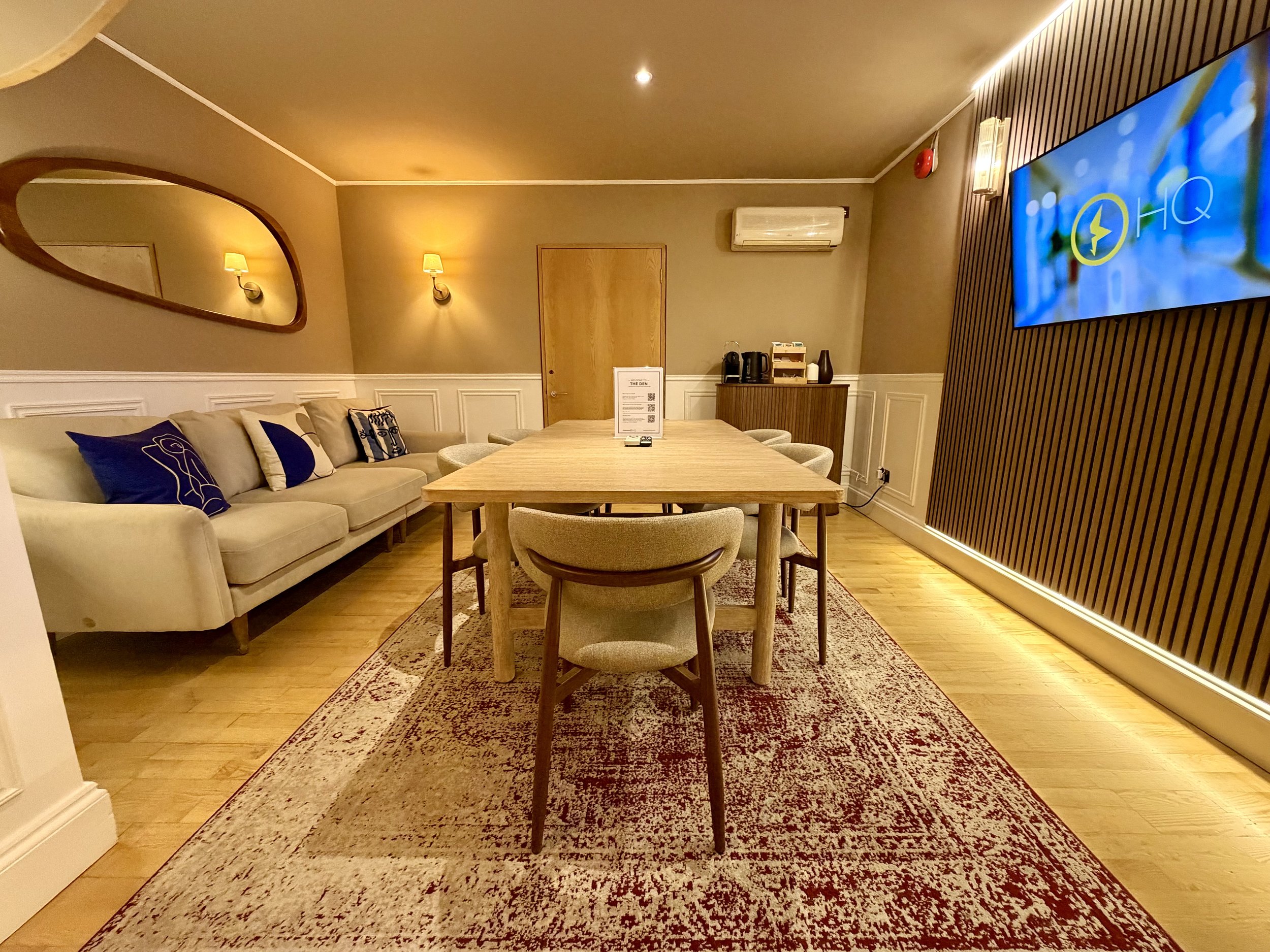 A boardroom with brown overtones, featuring a large oak table with chairs all around, a large TV attached to the wall and a generous sofa.