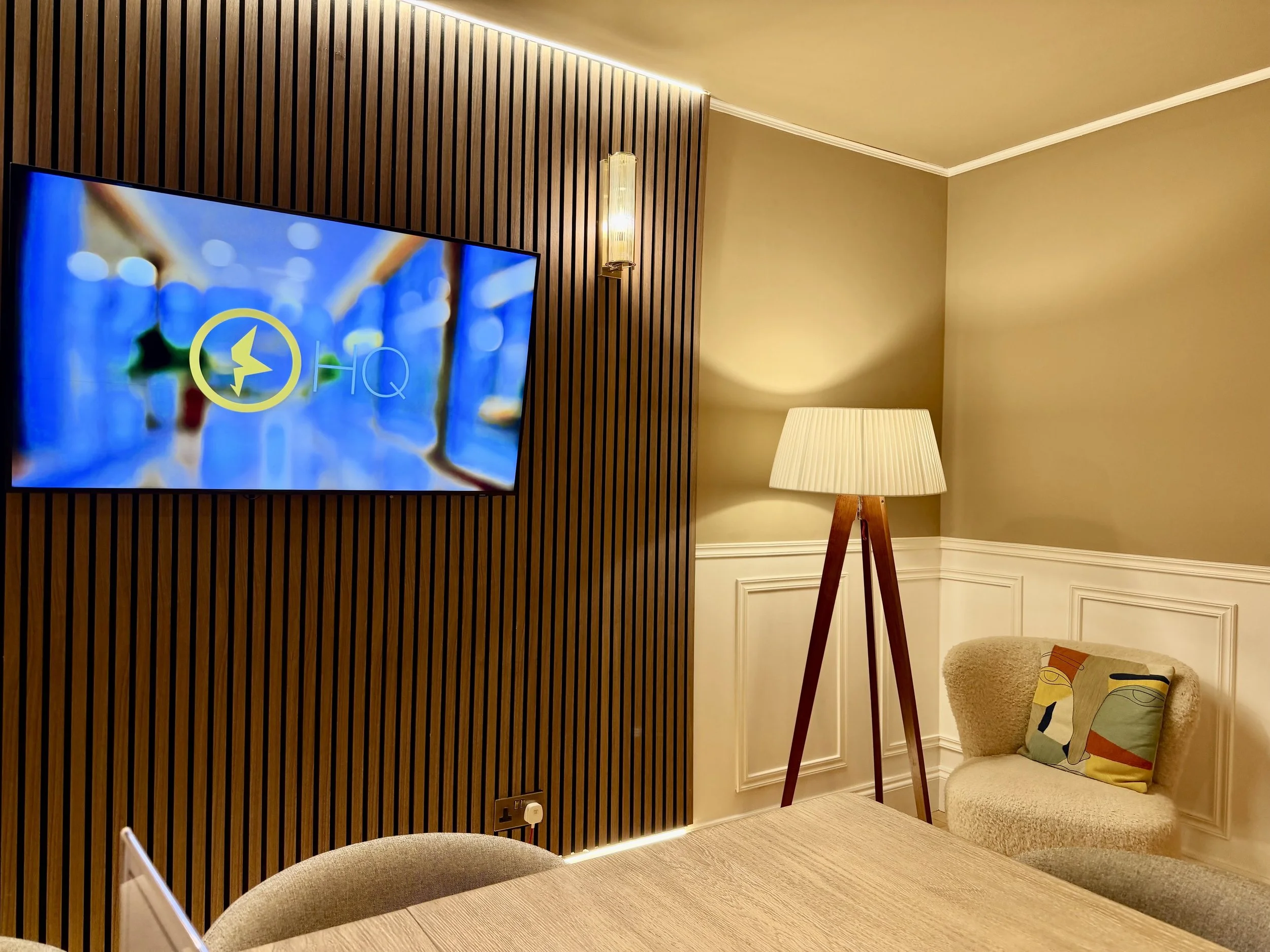 A big TV screen hangs on a feature wall with a large lamp and armchair nearby. In the front of shot, the edge of a large oak boardroom table and chairs.