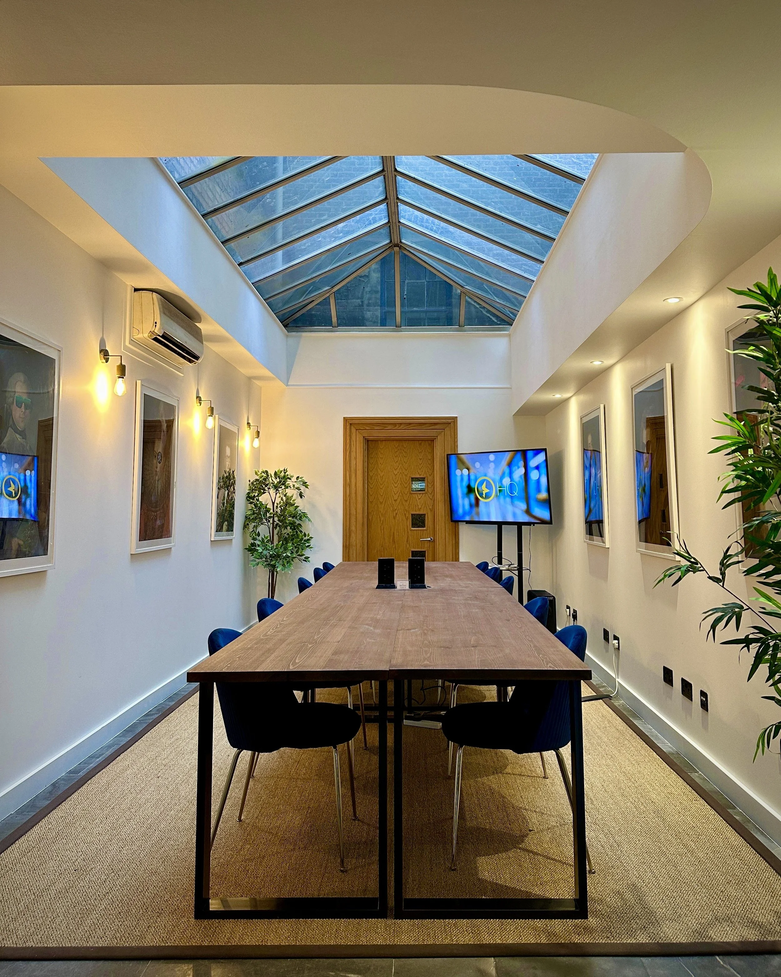 A large meeting room featuring a conservatory-style ceiling, with bright white walls and a large wooden-style table surrounded by blue chairs and large wooden door to the rear.