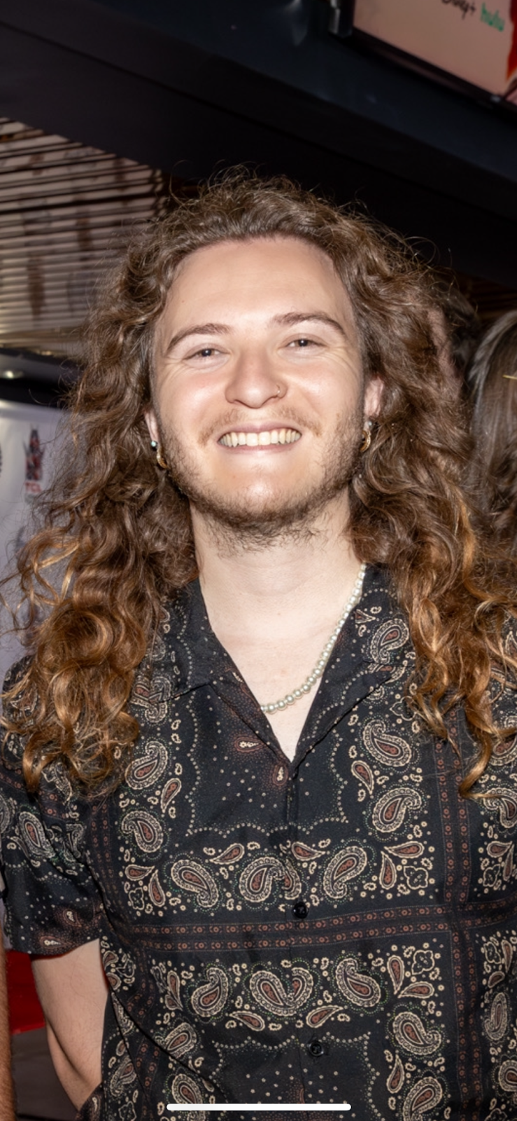 A smiling man with long, curly hair, wearing earrings, a beaded necklace, and a patterned black shirt, indoors at a social event.