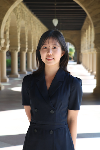 Young woman with shoulder-length dark hair standing under an archway in a courtyard, wearing a navy dress with buttons, smiling at the camera.