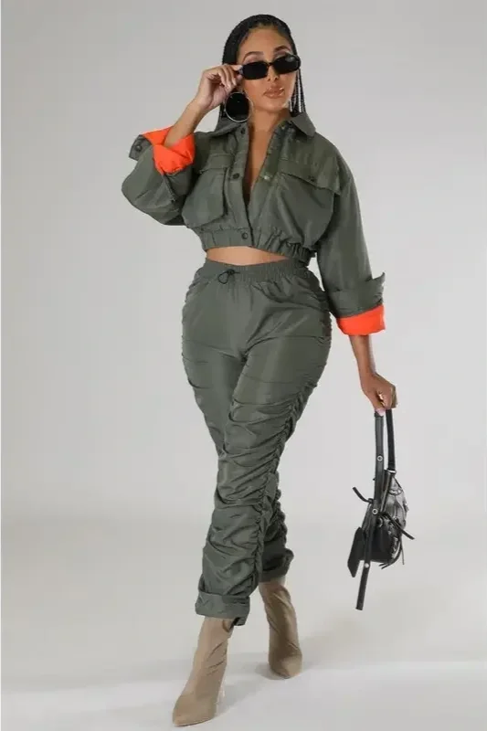 Woman wearing green cropped jacket with orange cuffs, matching green pants, beige boots, black sunglasses, and large hoop earrings, holding a small black handbag.