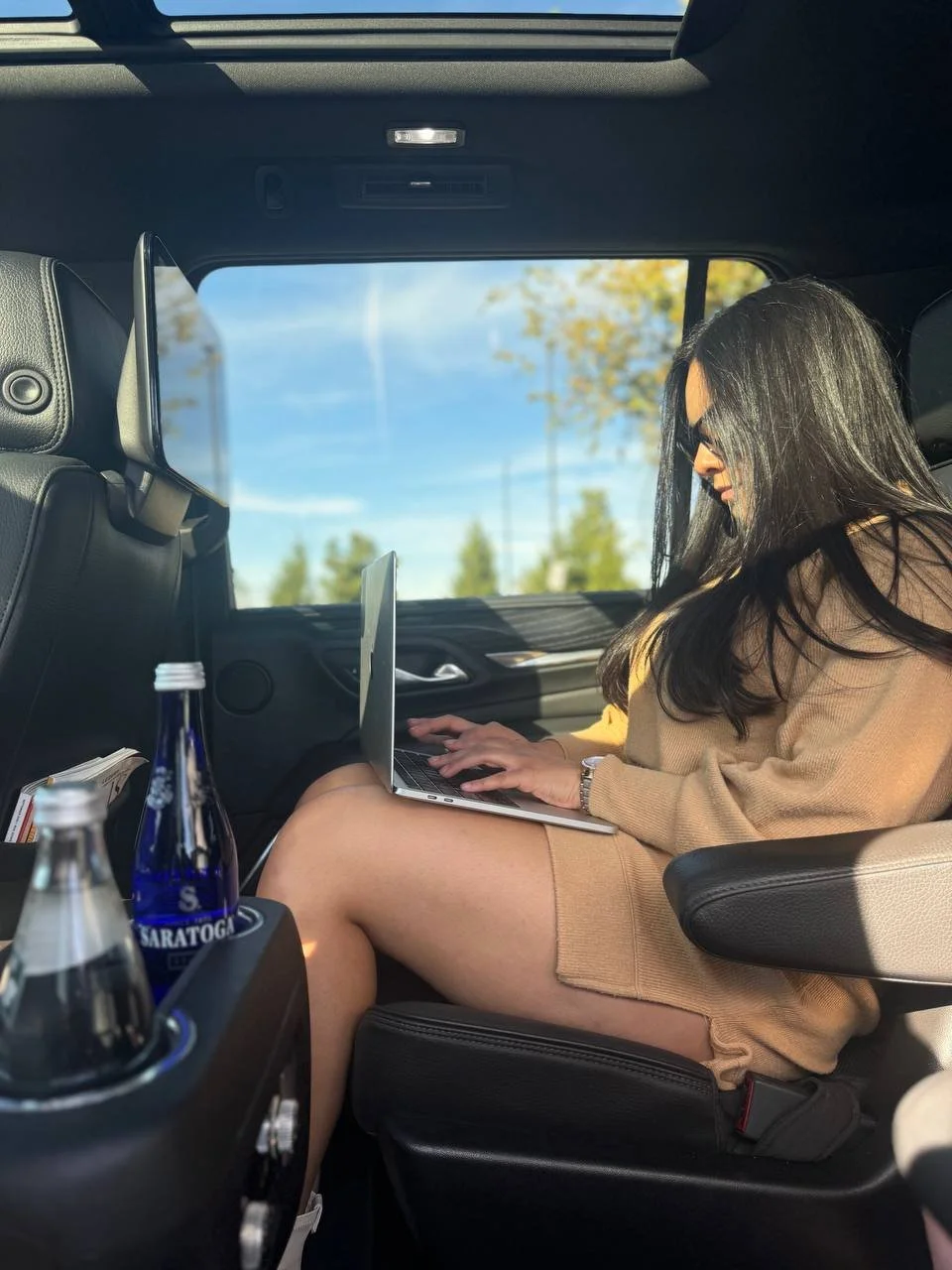 A woman with long black hair and sunglasses sitting in the back seat of a vehicle, working on a laptop. There are bottles of soda in the cup holder in front of her, and trees are visible outside the window.