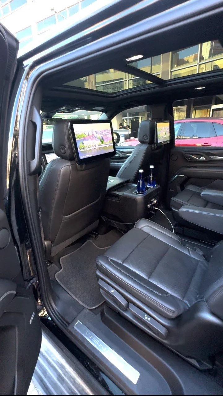 Interior of a luxury vehicle showing black leather seats, built-in screens on the back of front seats, and bottles of blue beverages in cup holders, with a sunroof overhead.