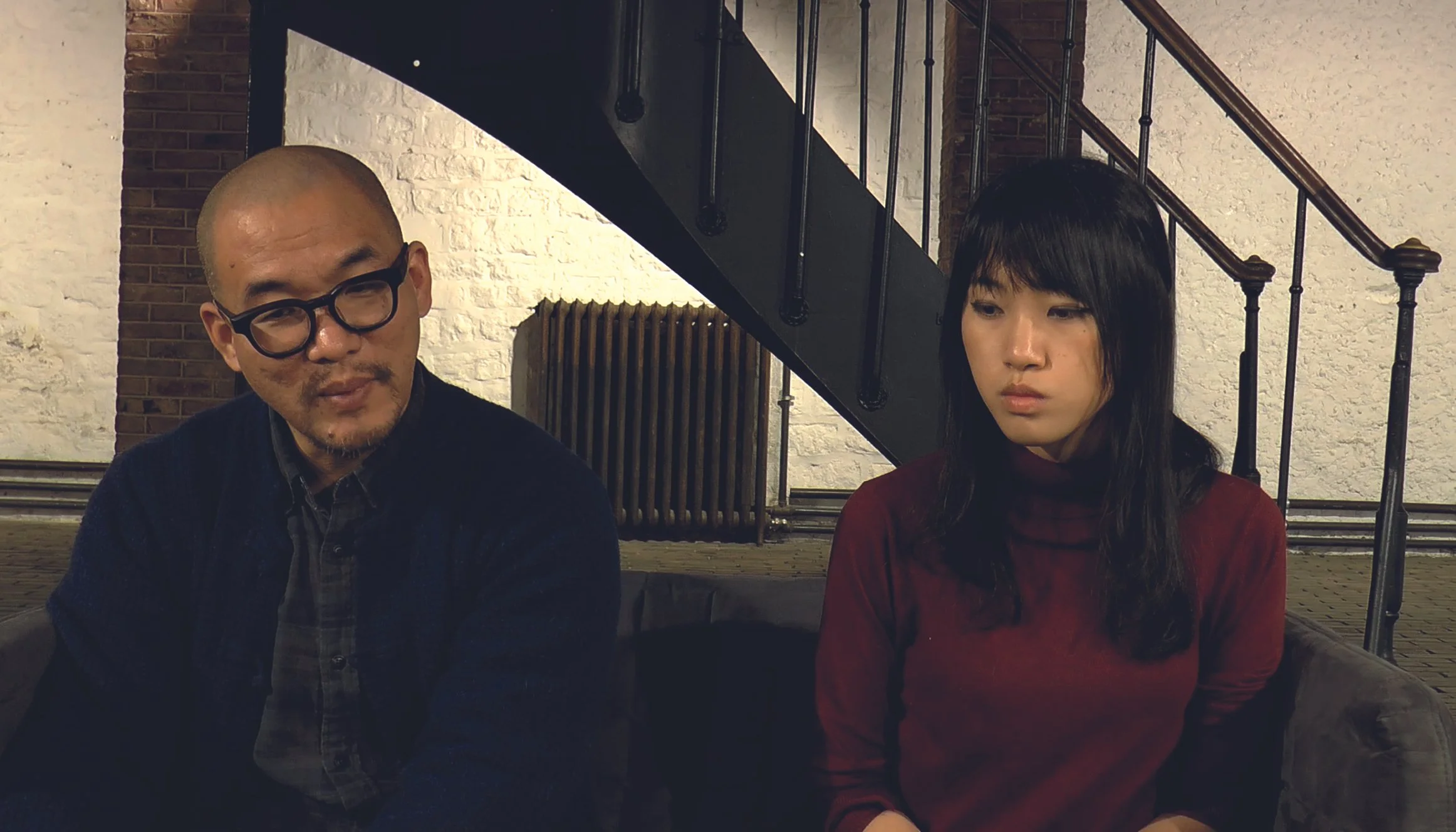 Un homme portant des lunettes noires et une chemise noire discute avec une femme aux cheveux noirs et à la chemise rouge dans un endroit intérieur avec un escalier en métal en arrière-plan.