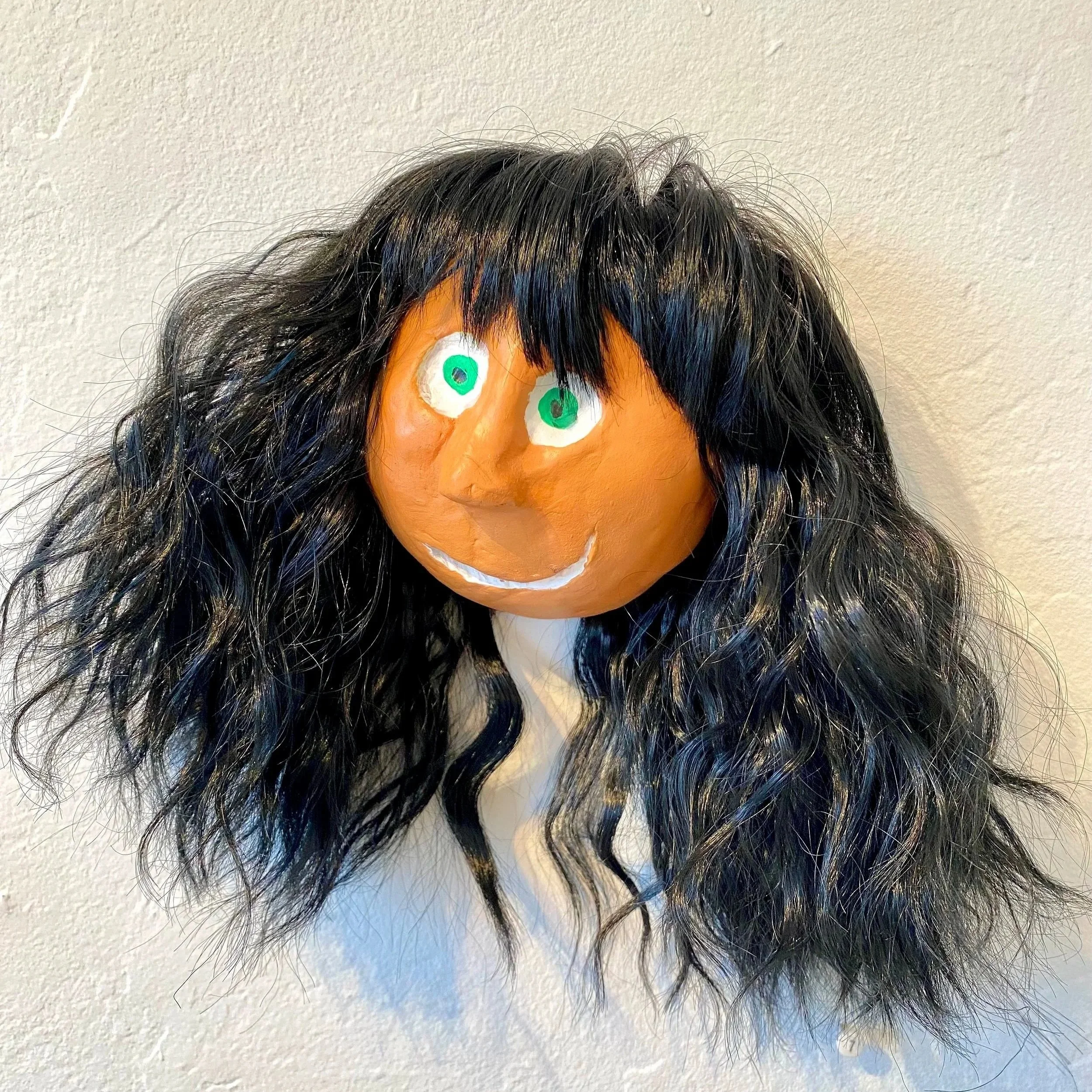 A human face mask with long black hair, bright green eyes, a big nose, and a big white smile, mounted on a wall.