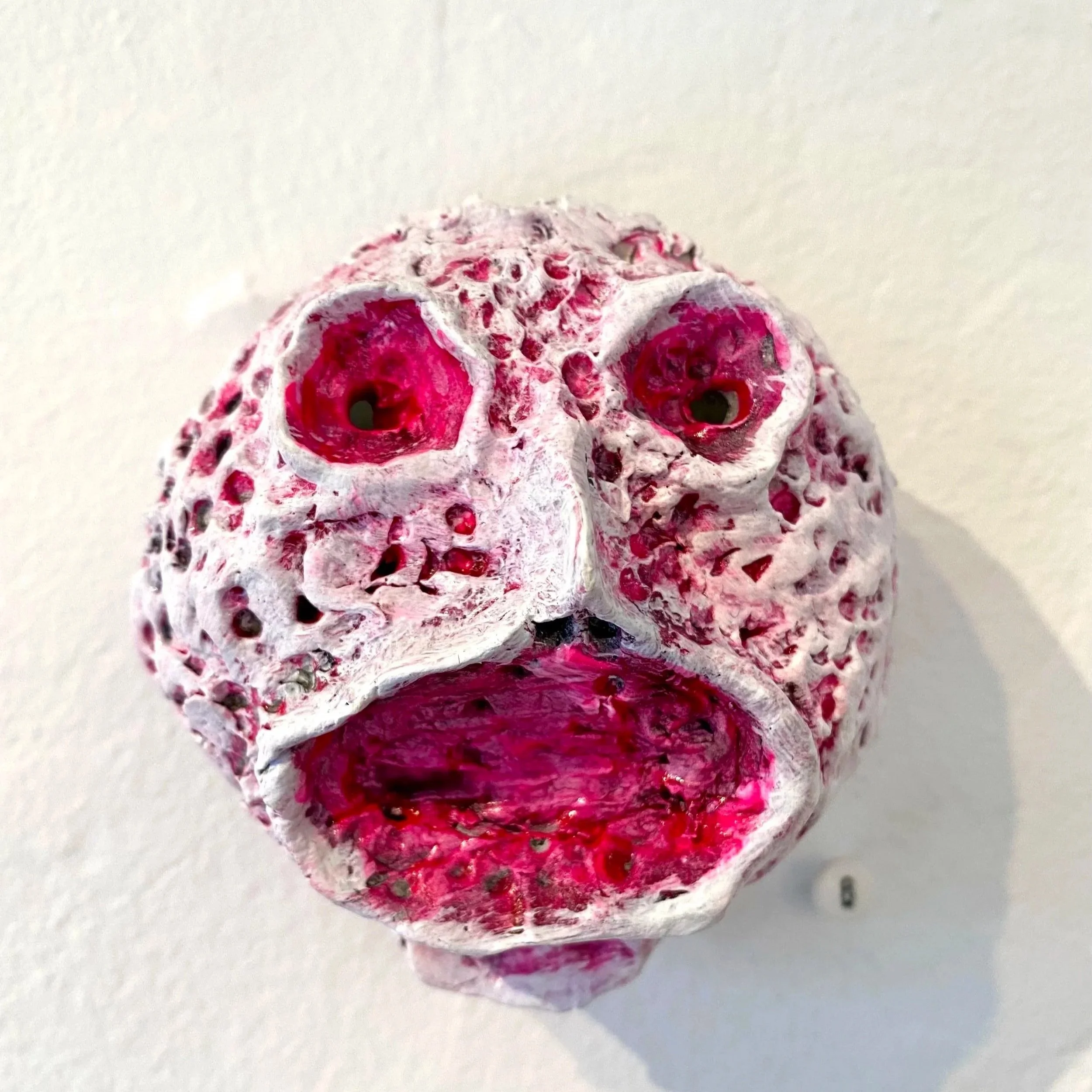 Sculpture of a face with hollow eyes and an open mouth, made from a porous material with a rough surface, painted white with pink accents.