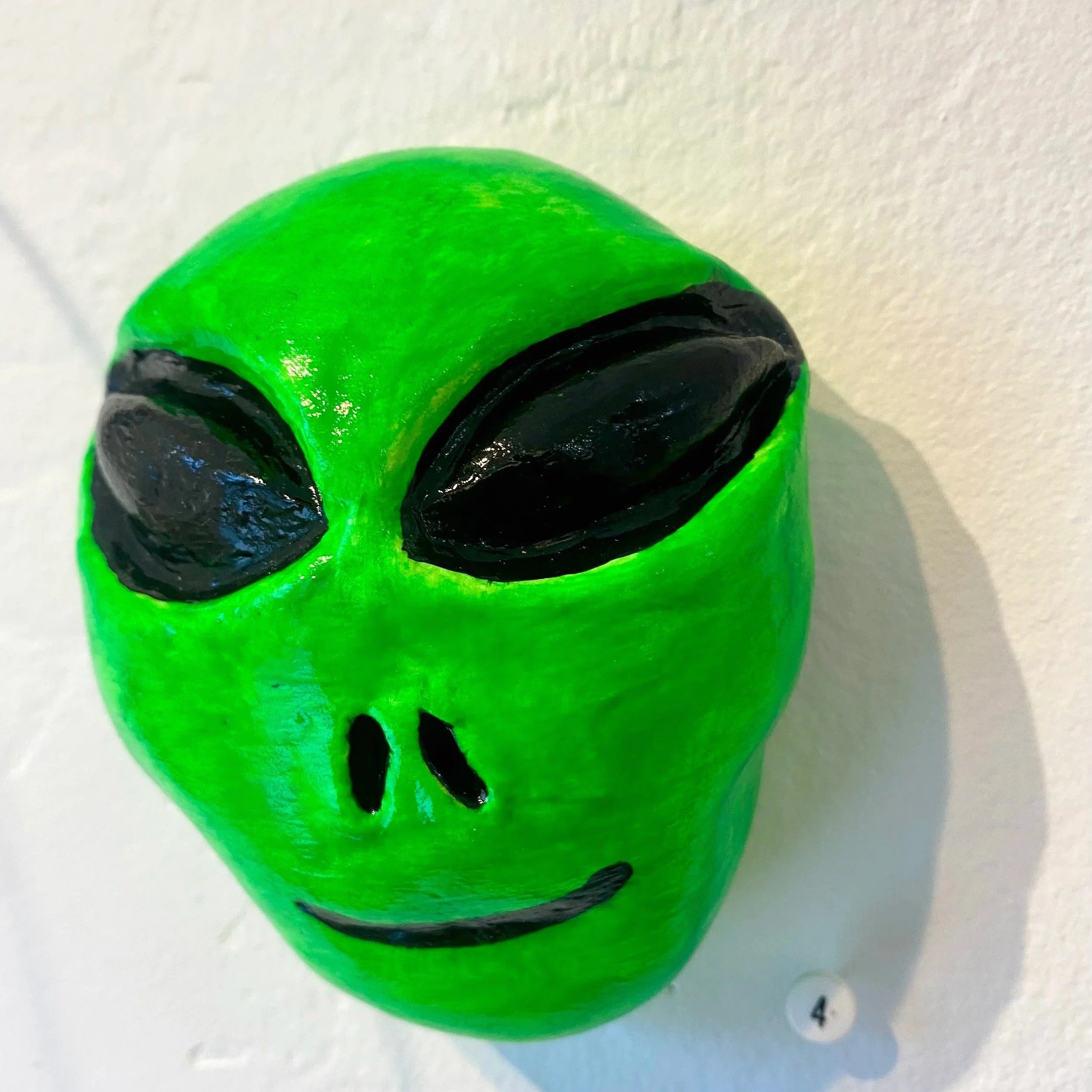 Green alien head sculpture with black eyes, nose, and mouth, mounted on a wall.