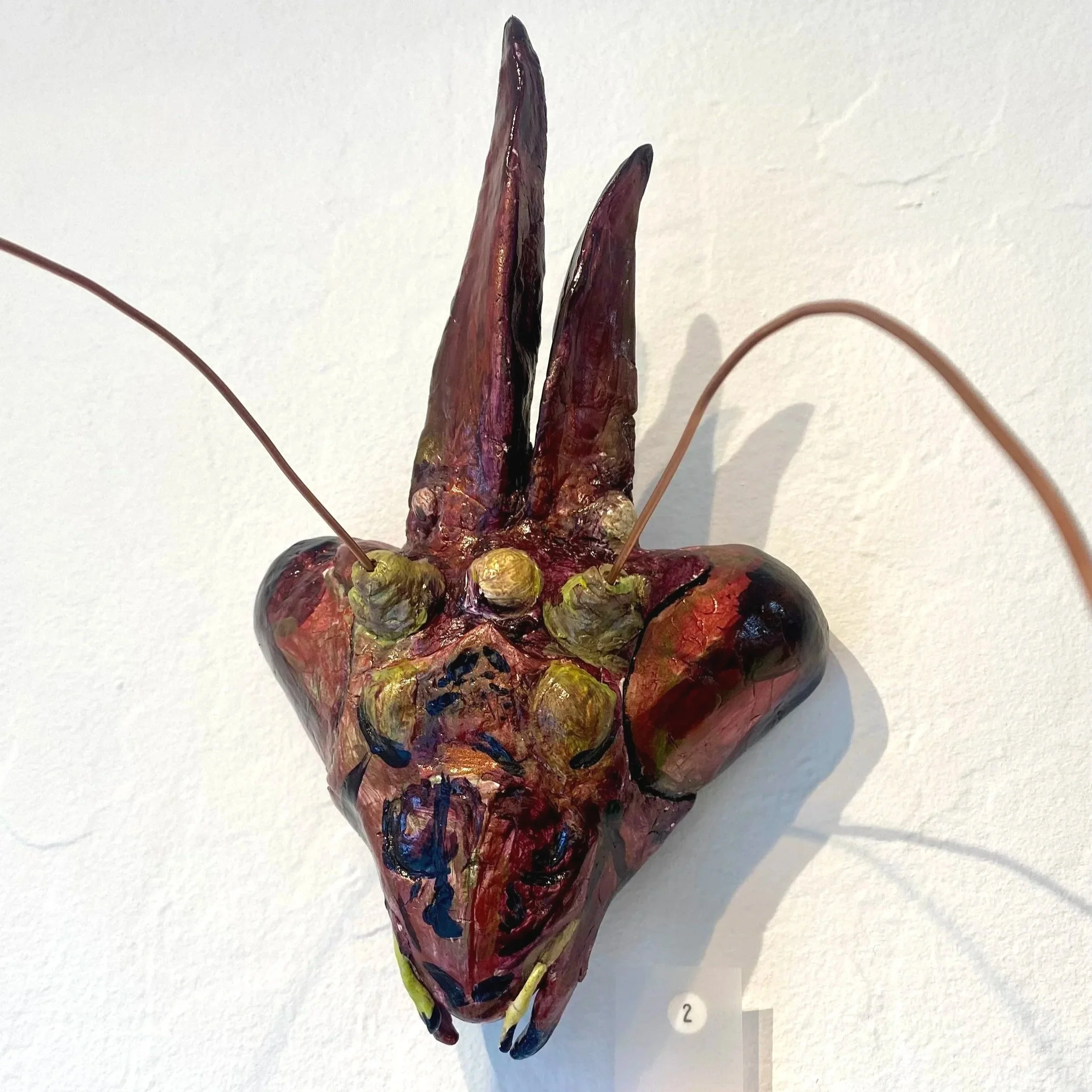 A colorful, abstract sculpture resembling a stylized animal face, with elongated ears, large eyes, and a pointed nose, mounted on a white wall.