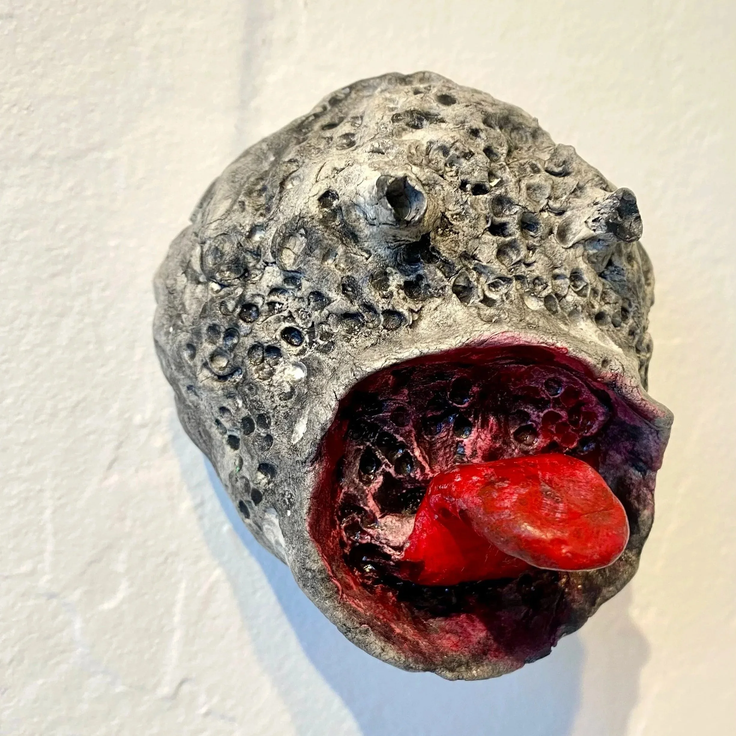 A textured sculpture resembling a face with a rock-like surface, an open mouth with a red tongue, and a dark interior against a plain wall.