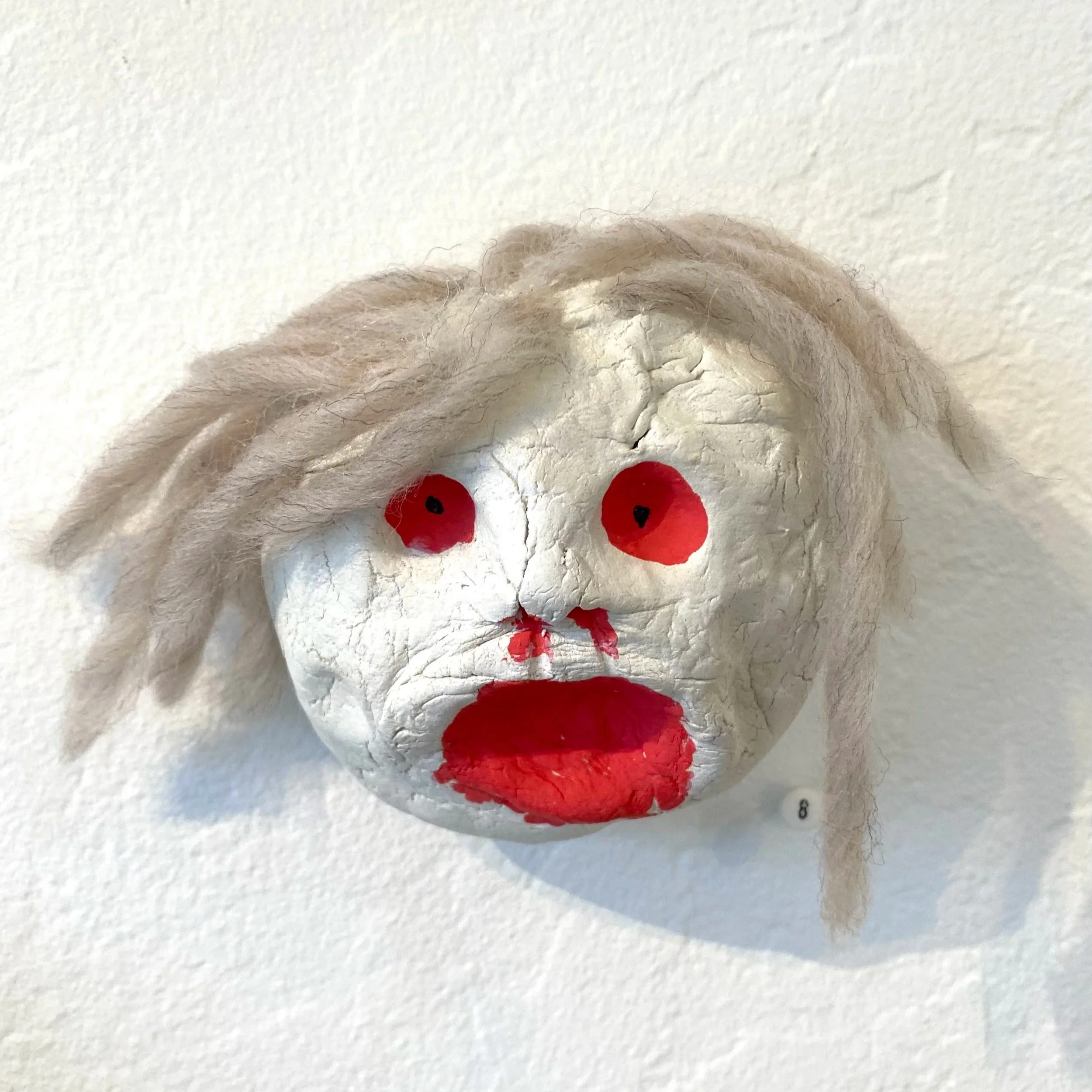 A face sculpture made of cracked white material with red painted eyes and mouth, and light-colored hair made of felt or yarn, mounted on a white wall.