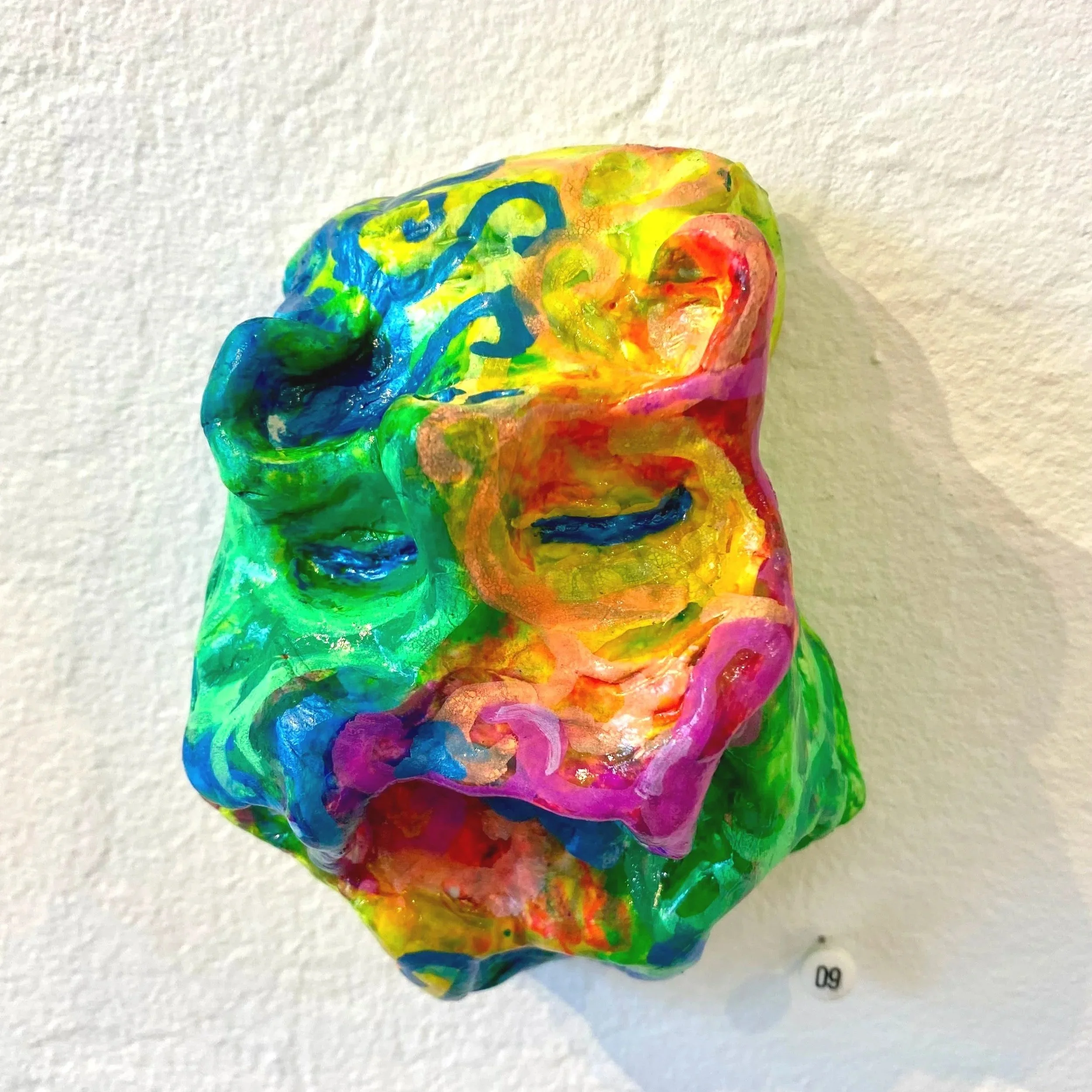 Colorful abstract sculpture resembling a distorted face with various vibrant colors including blue, green, yellow, red, and pink, mounted on a white wall.