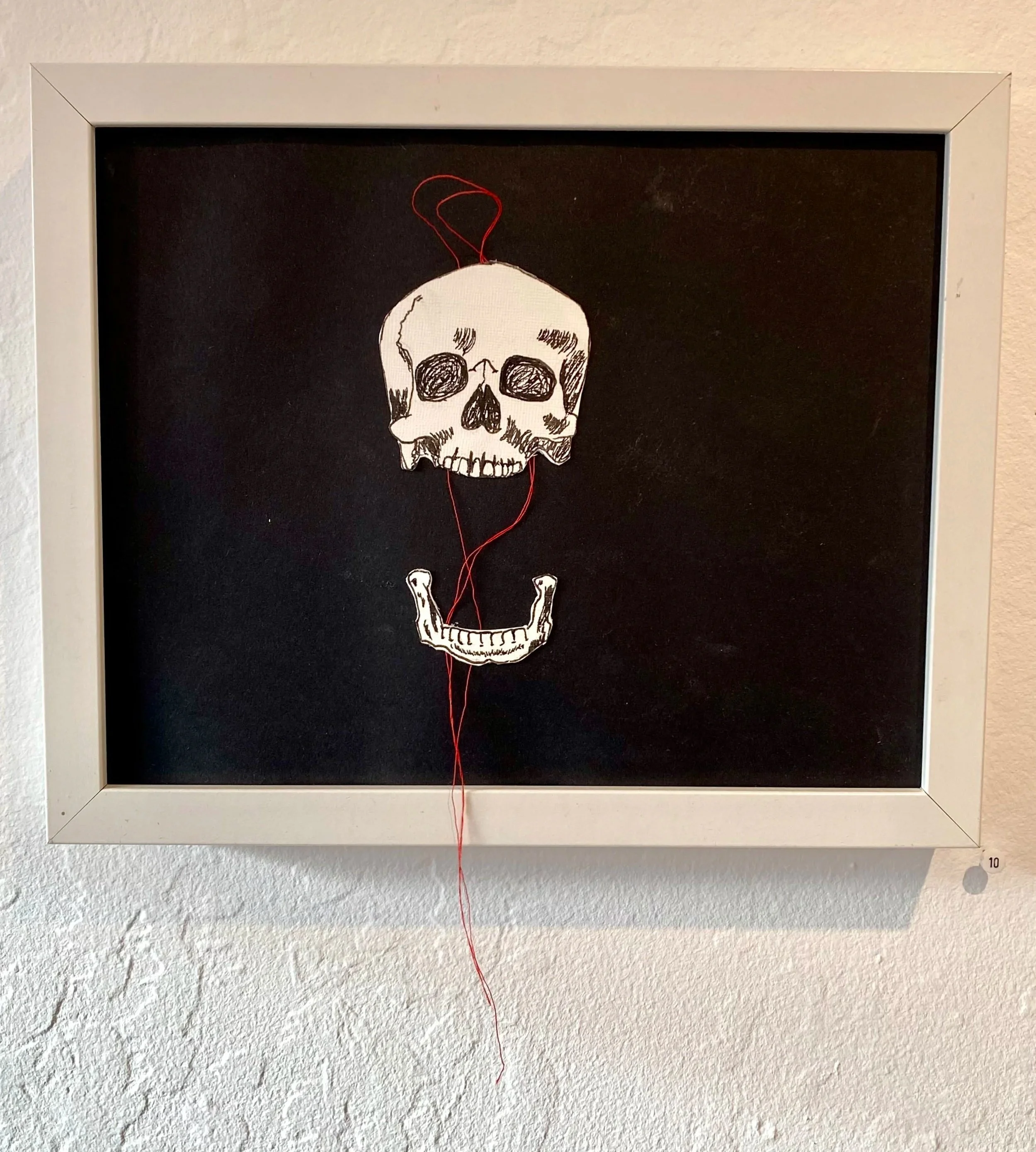 Artwork featuring a skull with hollow eyes and an open mouth, with a stitched grin, depicted on black background. A red thread is attached to the skull, hanging downward.