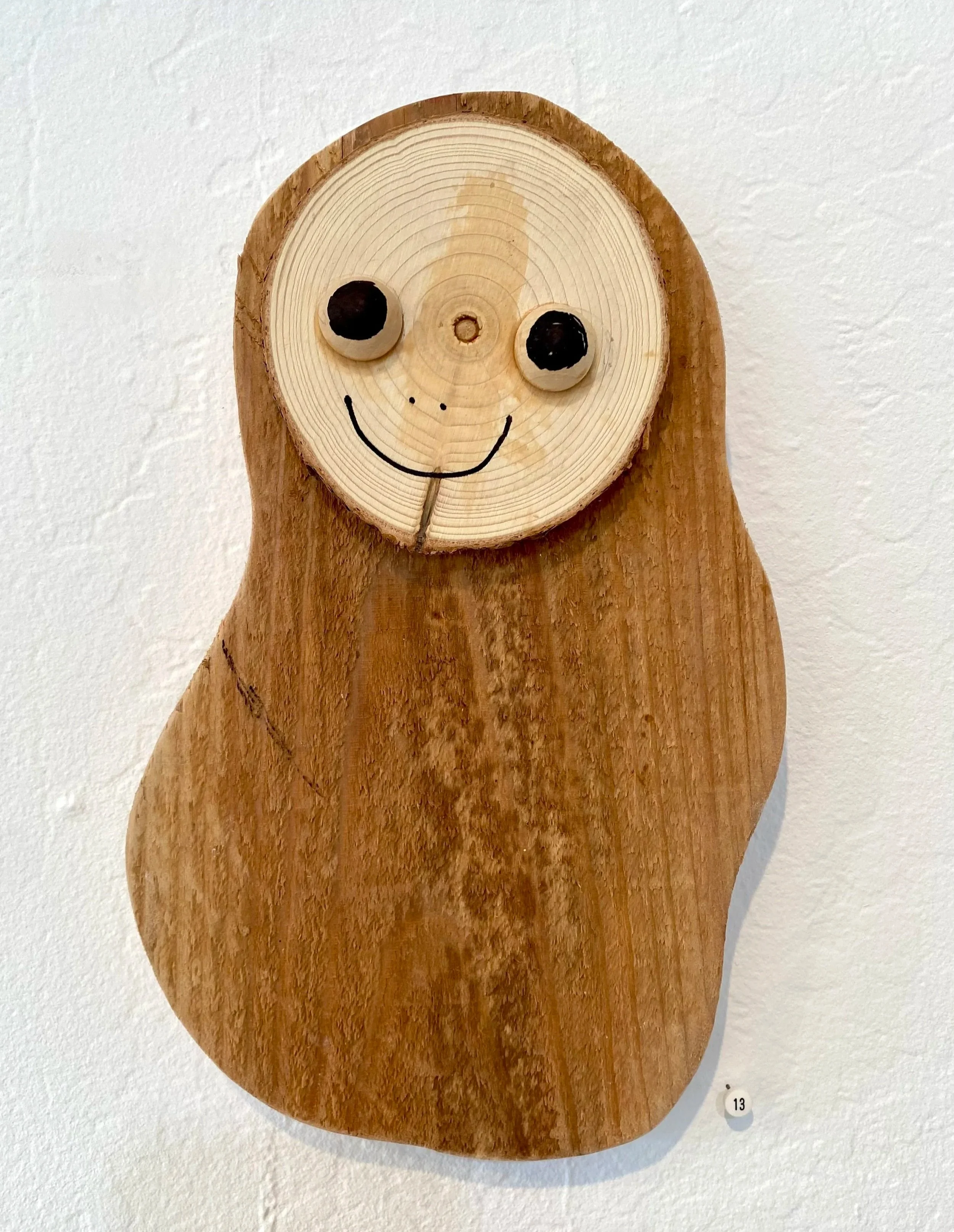 Wooden art piece resembling a smiling animal, with a circular light-colored wood face featuring black painted eyes, a simple black painted smile, and small dots for a nose, mounted on a larger irregular dark wood body.