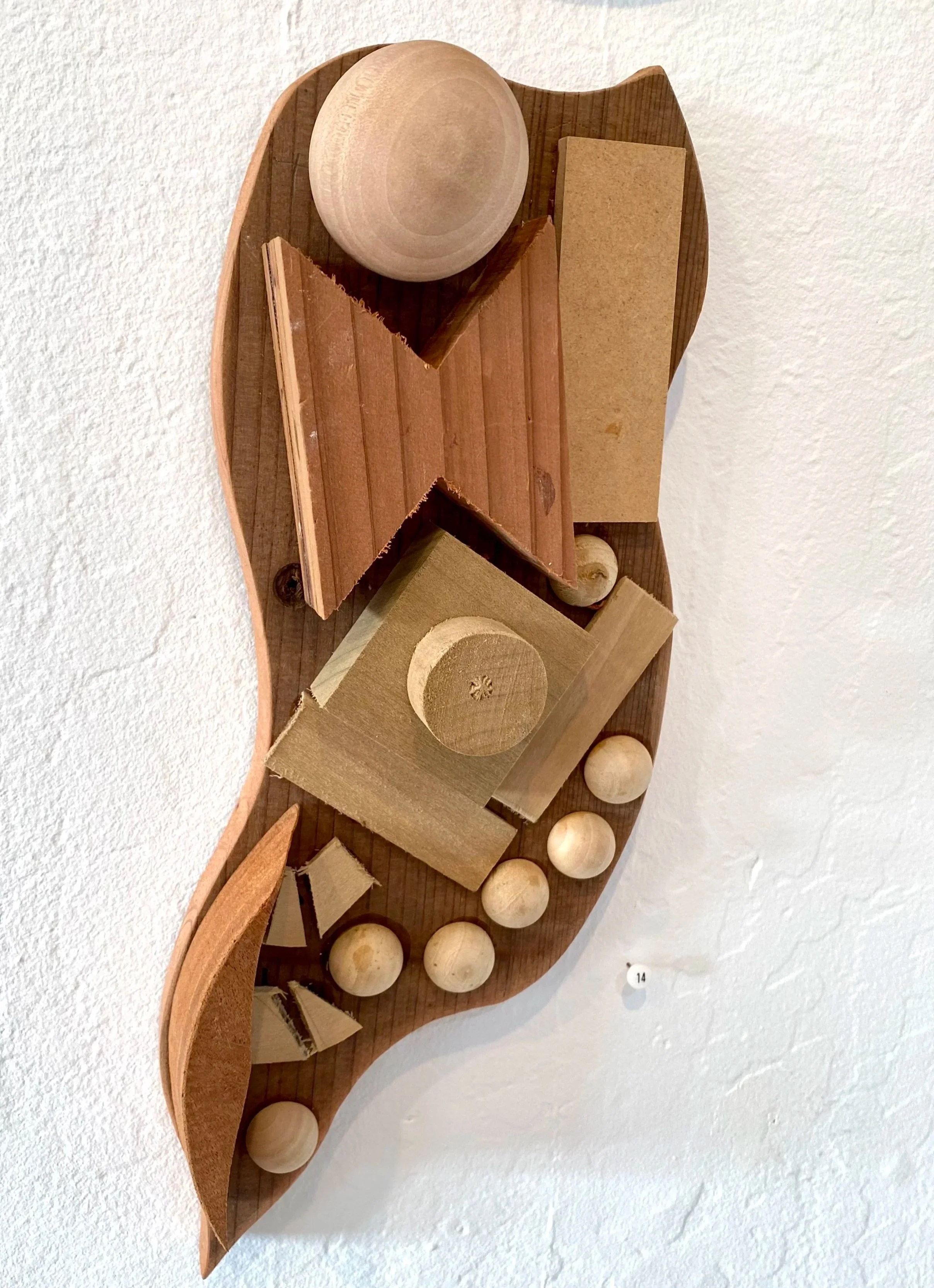 Abstract wooden sculpture with curved base, various geometric shapes, and small wooden balls mounted on a white wall.