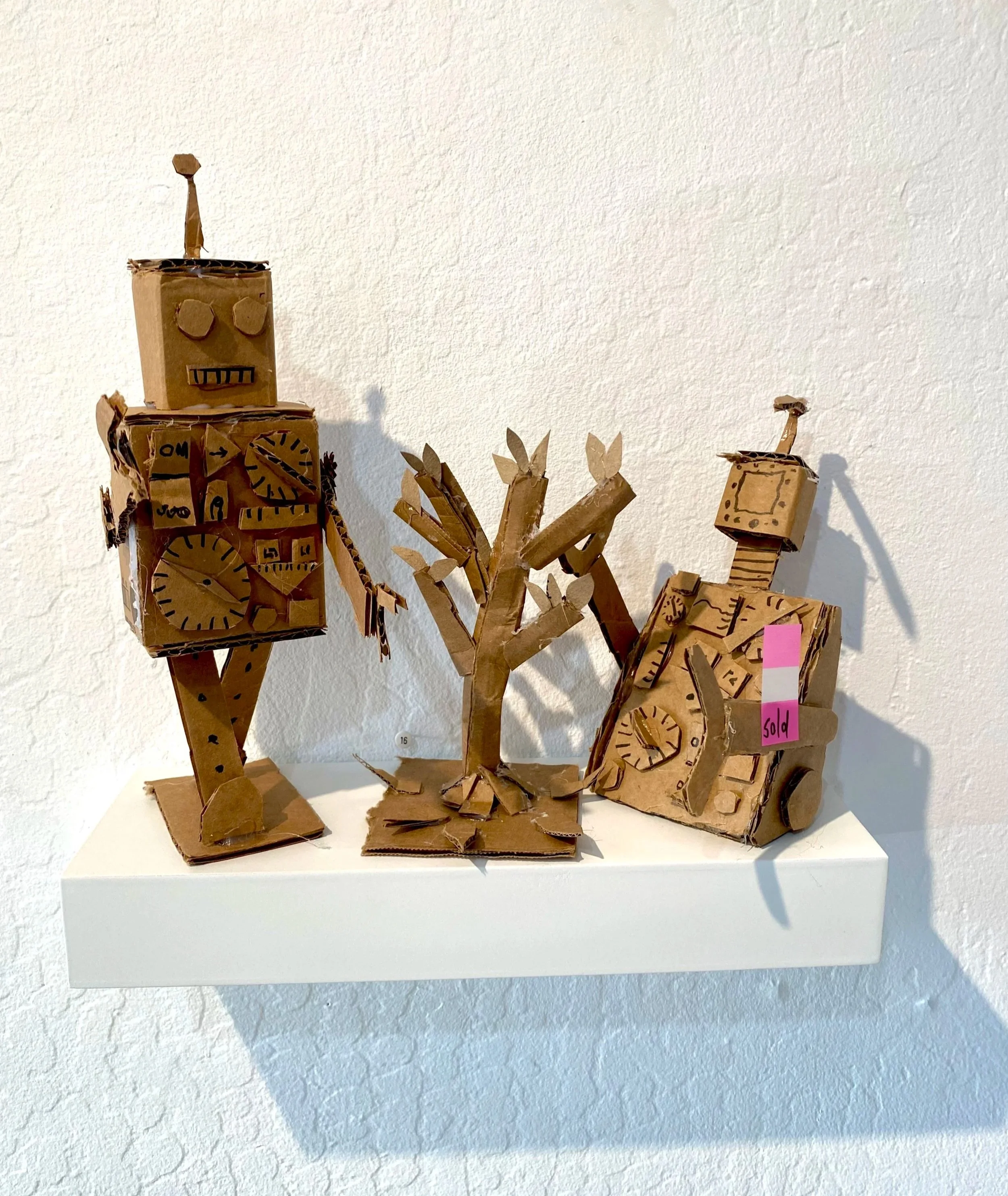 Two cardboard robot sculptures on a white shelf with a cardboard tree between them, all made from brown cardboard with hand-drawn details.
