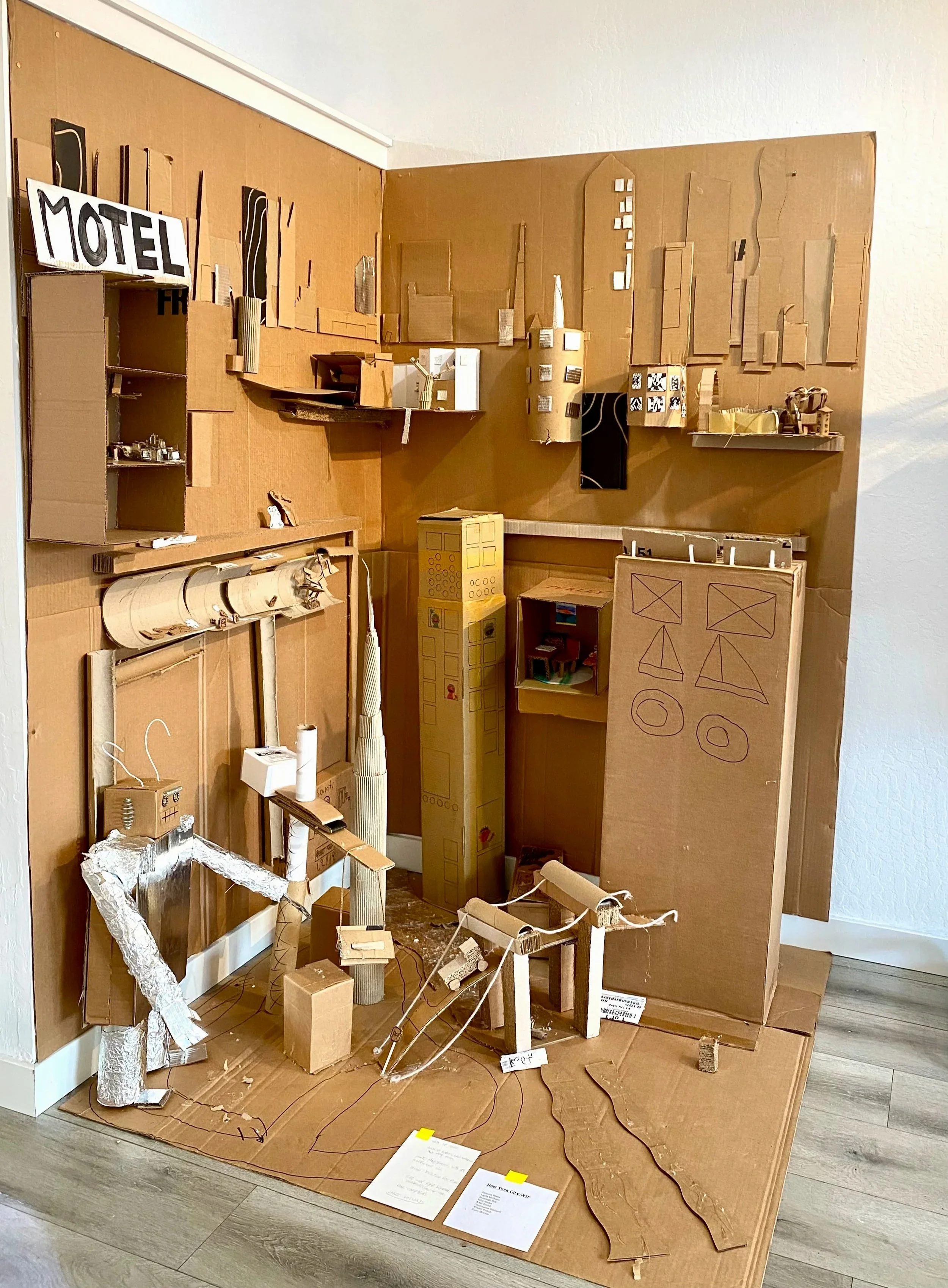 A detailed architectural model built from cardboard, featuring buildings, a motel sign, and urban layout elements.