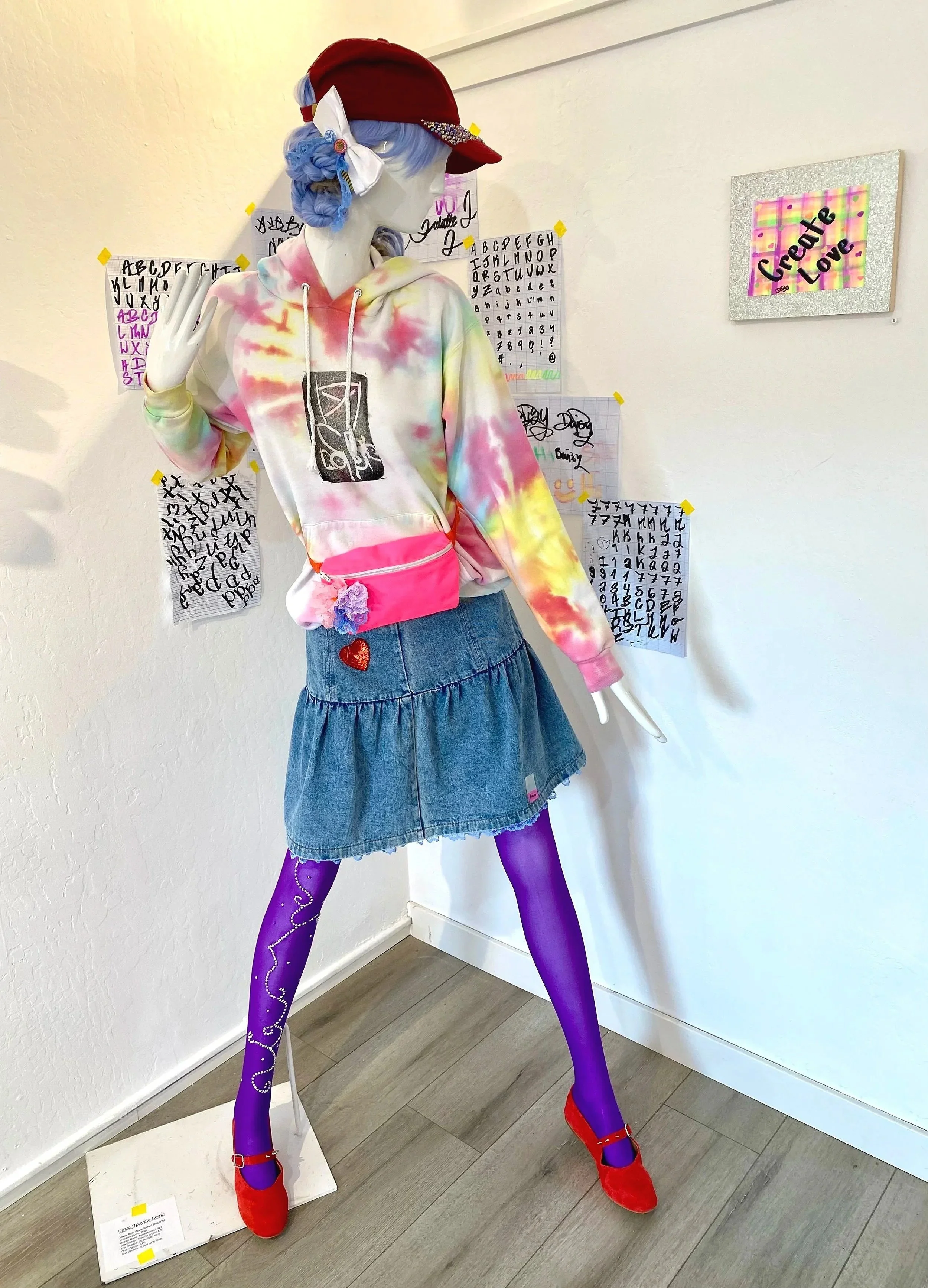 A mannequin dressed in colorful fashion is standing against a wall with various handwritten alphabet and doodle posters. The mannequin wears a tie-dye hoodie, a denim skirt, purple tights with doodles, red shoes, a red cap, and a large bow in blue ha