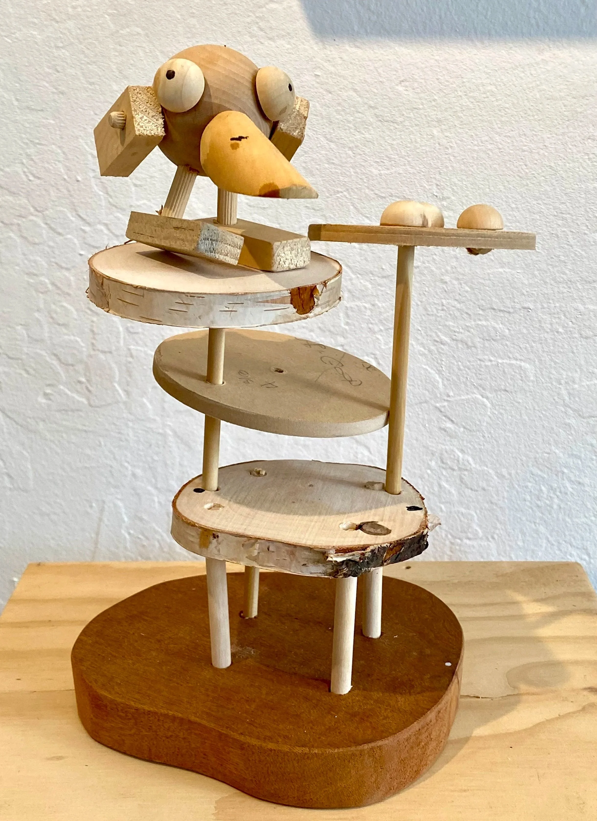 Wooden sculpture resembling a bird with a large beak, round eyes, and a head made of wooden blocks, standing on a multi-layered wooden base with various shapes and sizes of wooden discs and sticks.