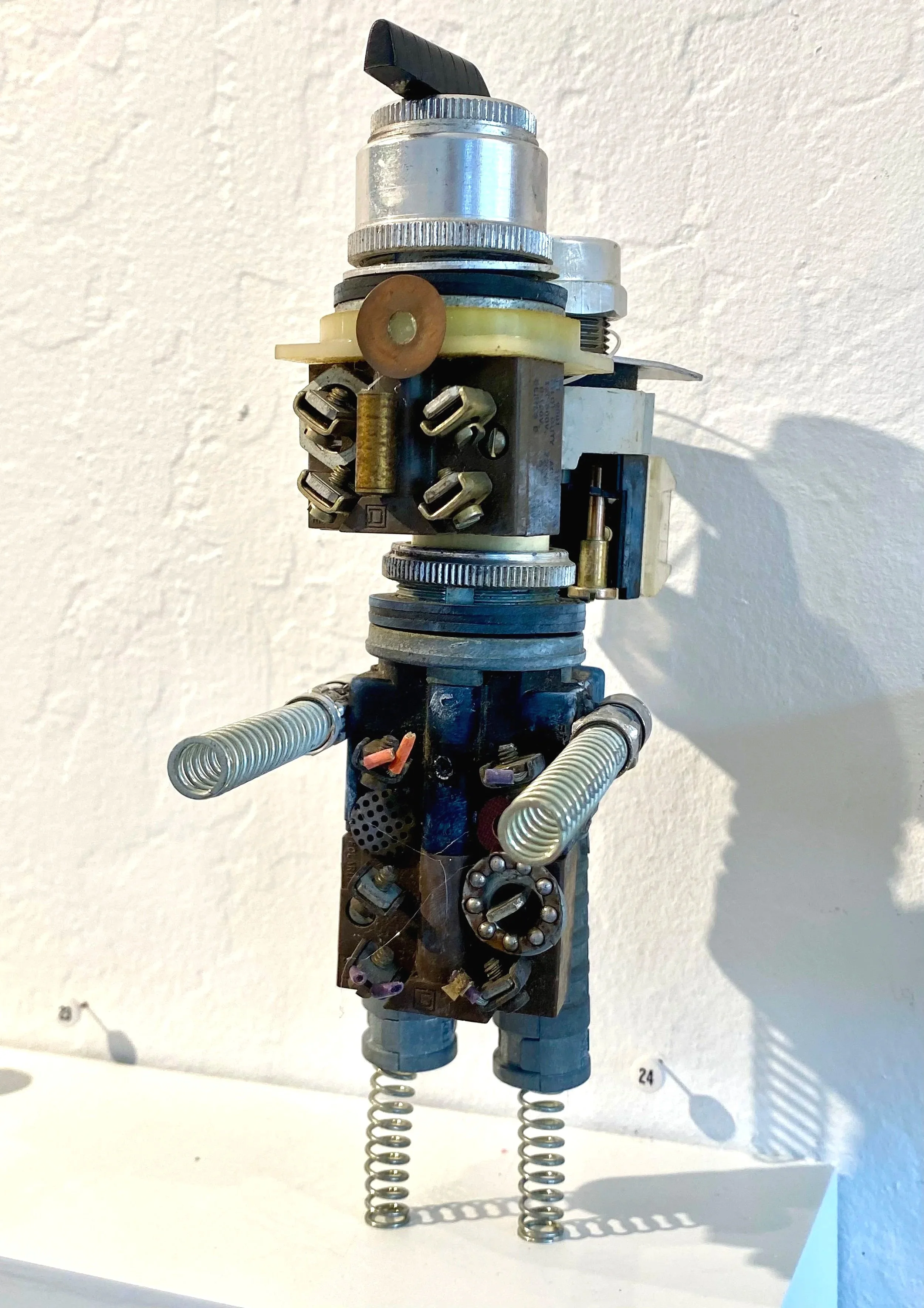 A humanoid figure made from various mechanical parts, with coiled wire arms and metal springs for legs, standing against a plain wall.