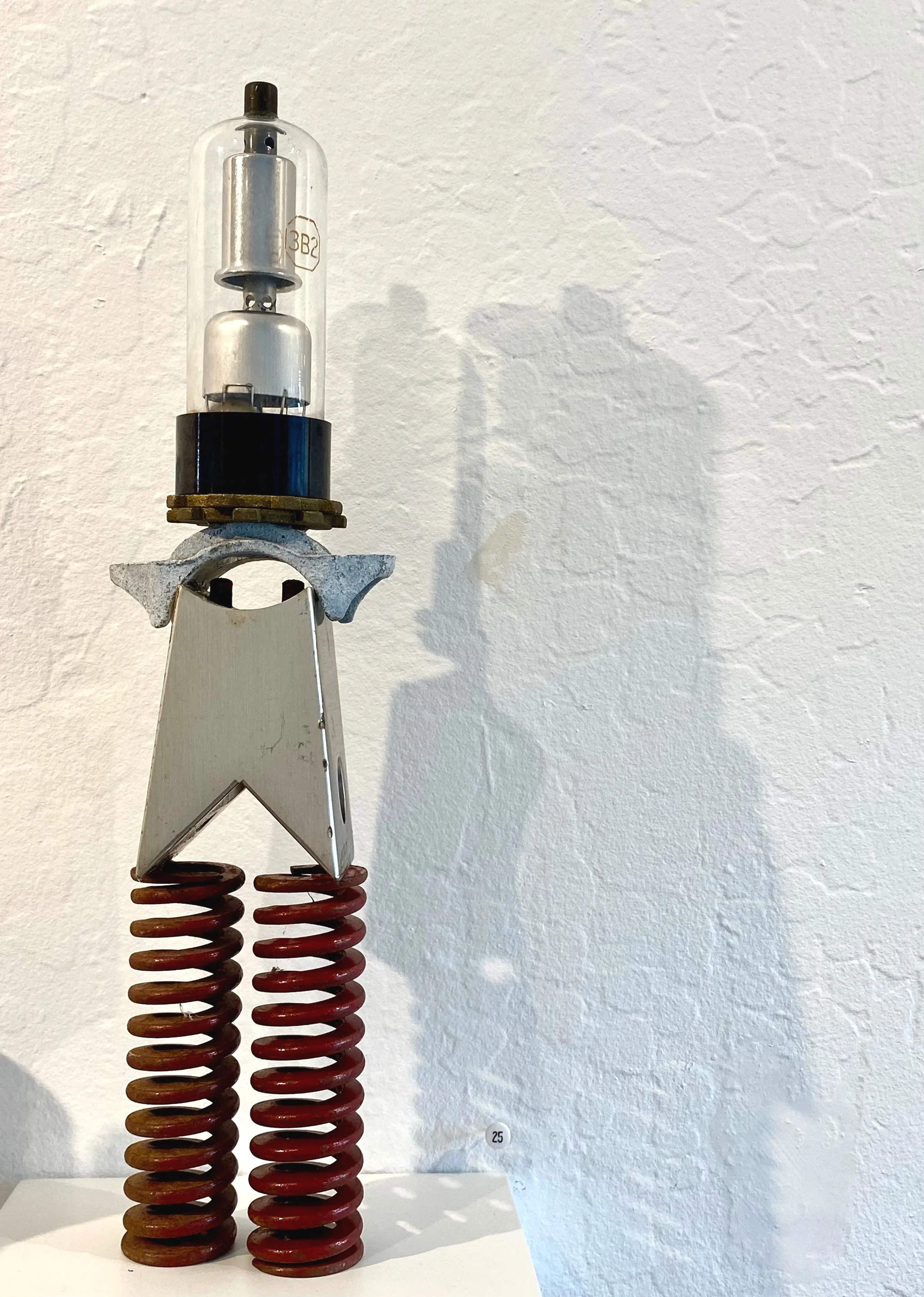 An art sculpture resembling a robot made from various metal parts, with two red coil springs for legs, a flat metallic body, and a glass vacuum tube on top.
