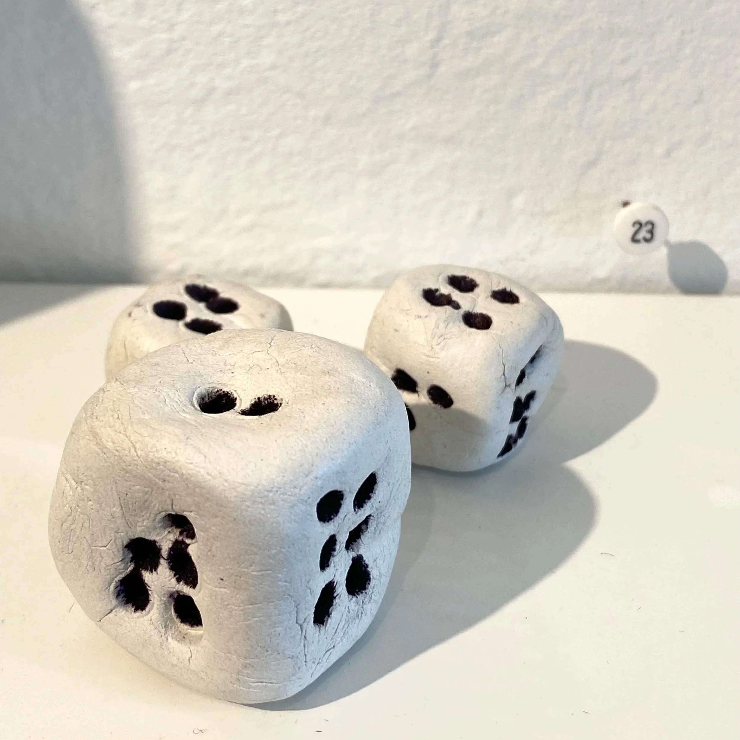 Three white ceramic dice with black dots on a white surface, with a small white ball labeled 23 in the background.