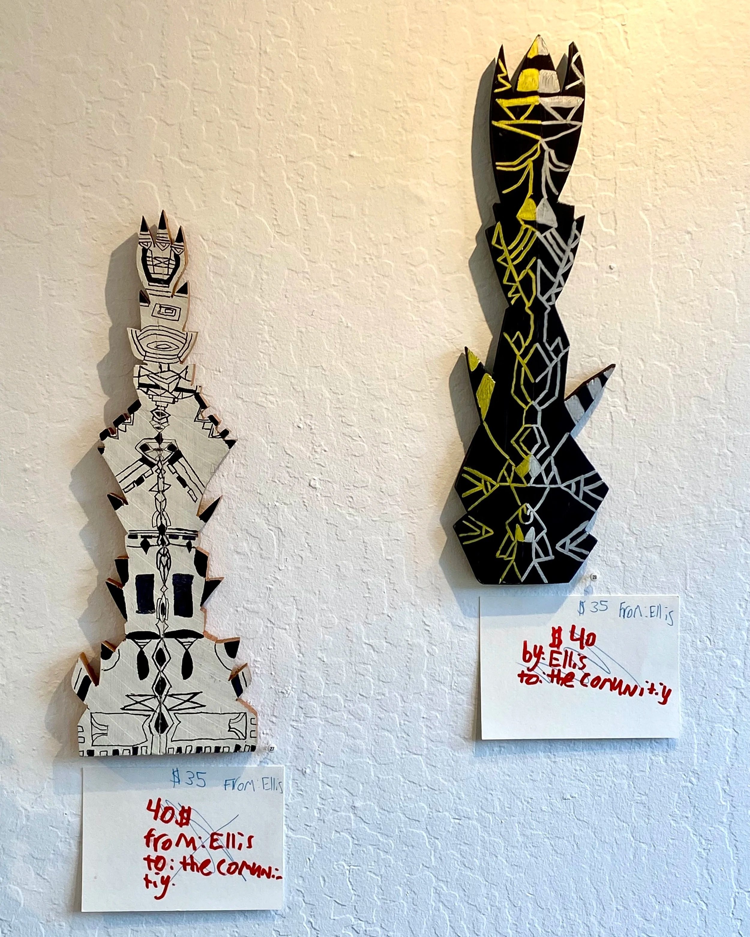 Two abstract black and white tribal-style wooden art pieces mounted on a white wall, each with a note below mentioning a price of 35 dollars and a message from Ellis to the community.
