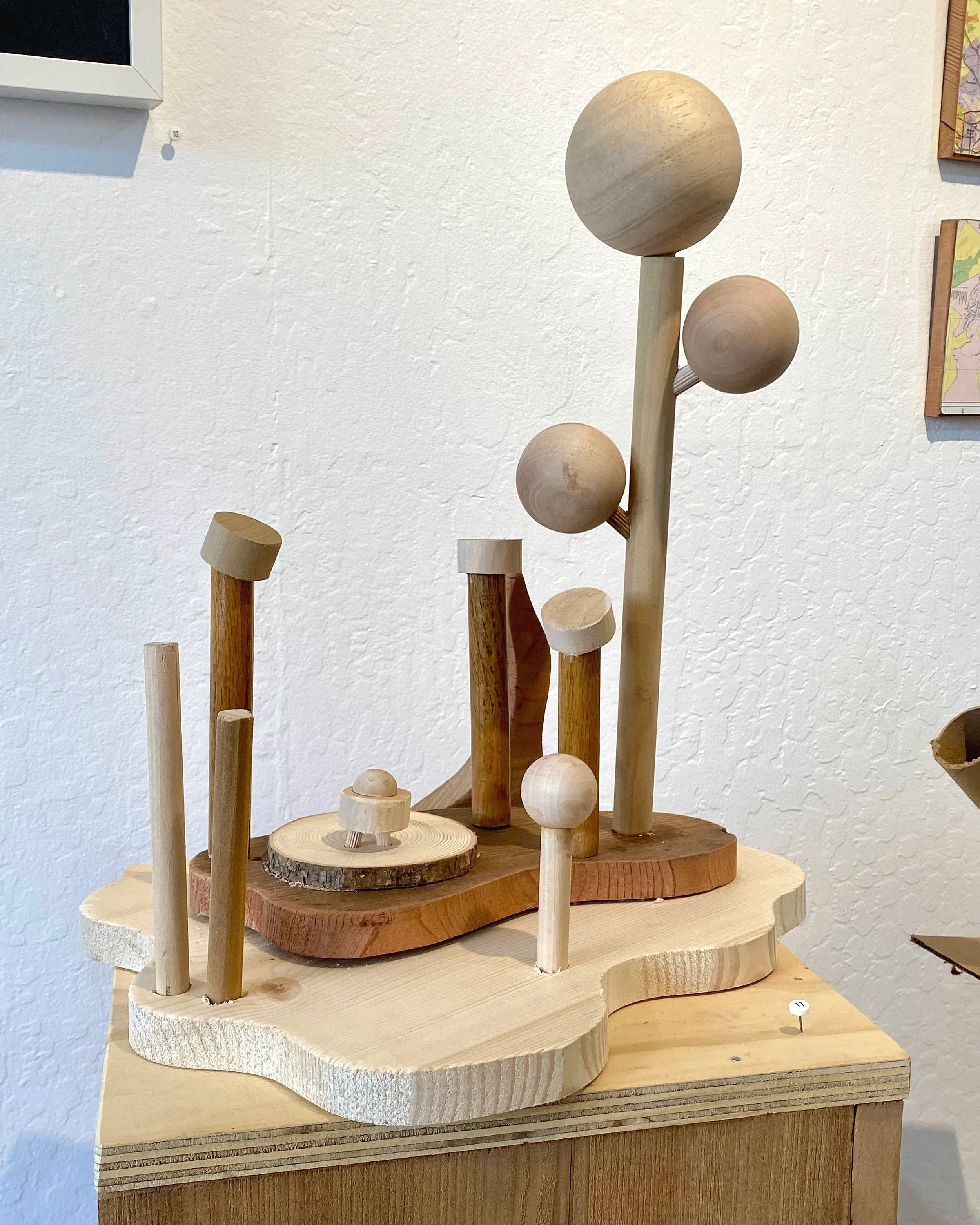 Wooden art sculpture with various vertical and horizontal wooden spheres and cylinders on a layered wooden base, displayed on a wooden stand against a white wall.