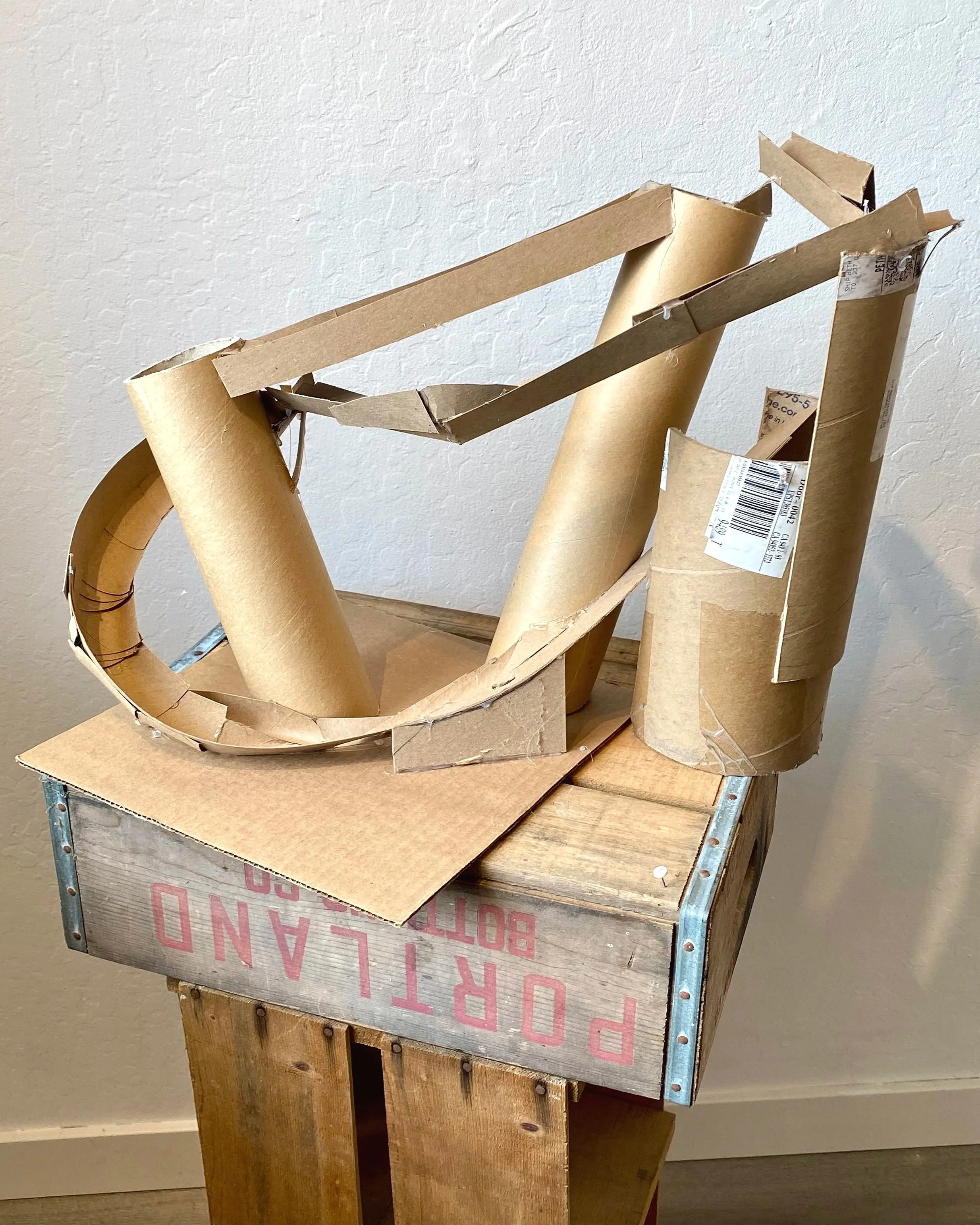 A sculpture made from cardboard tubes and pieces, assembled on a wooden crate with metal corner protectors. The sculpture has an abstract, curved structure with various angled and rolled cardboard parts.