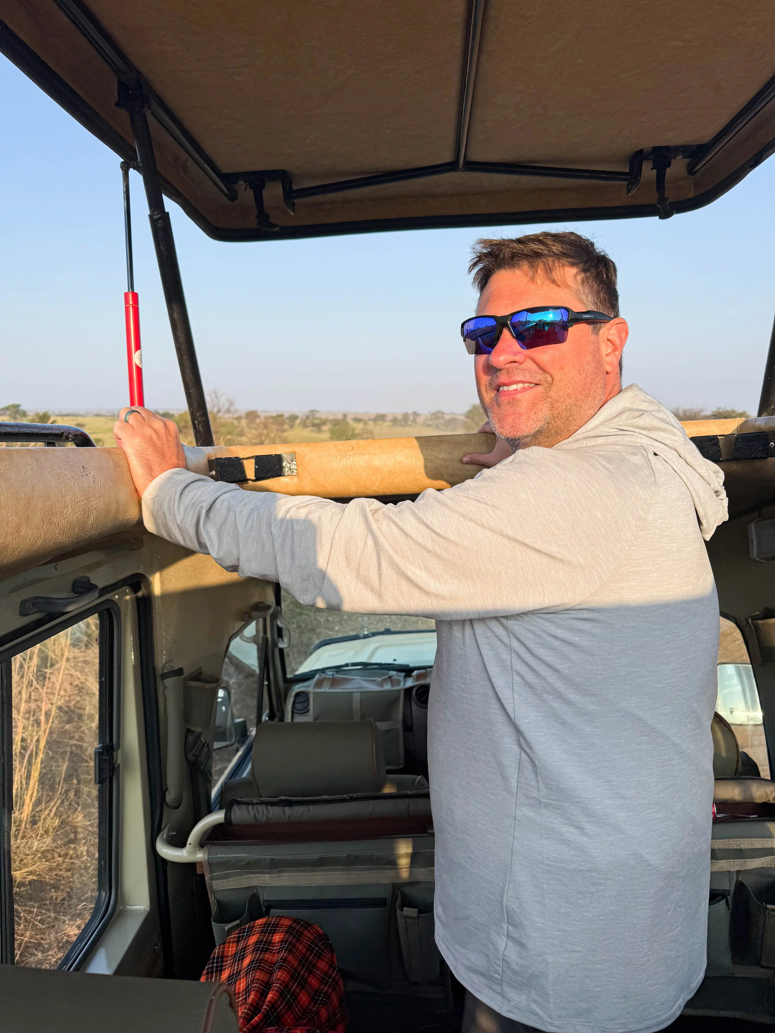 A man wearing sunglasses and a light grey hoodie stands inside a safari vehicle, holding onto the top frame and smiling at the camera. The vehicle is on a safari in an open landscape with a clear sky.
