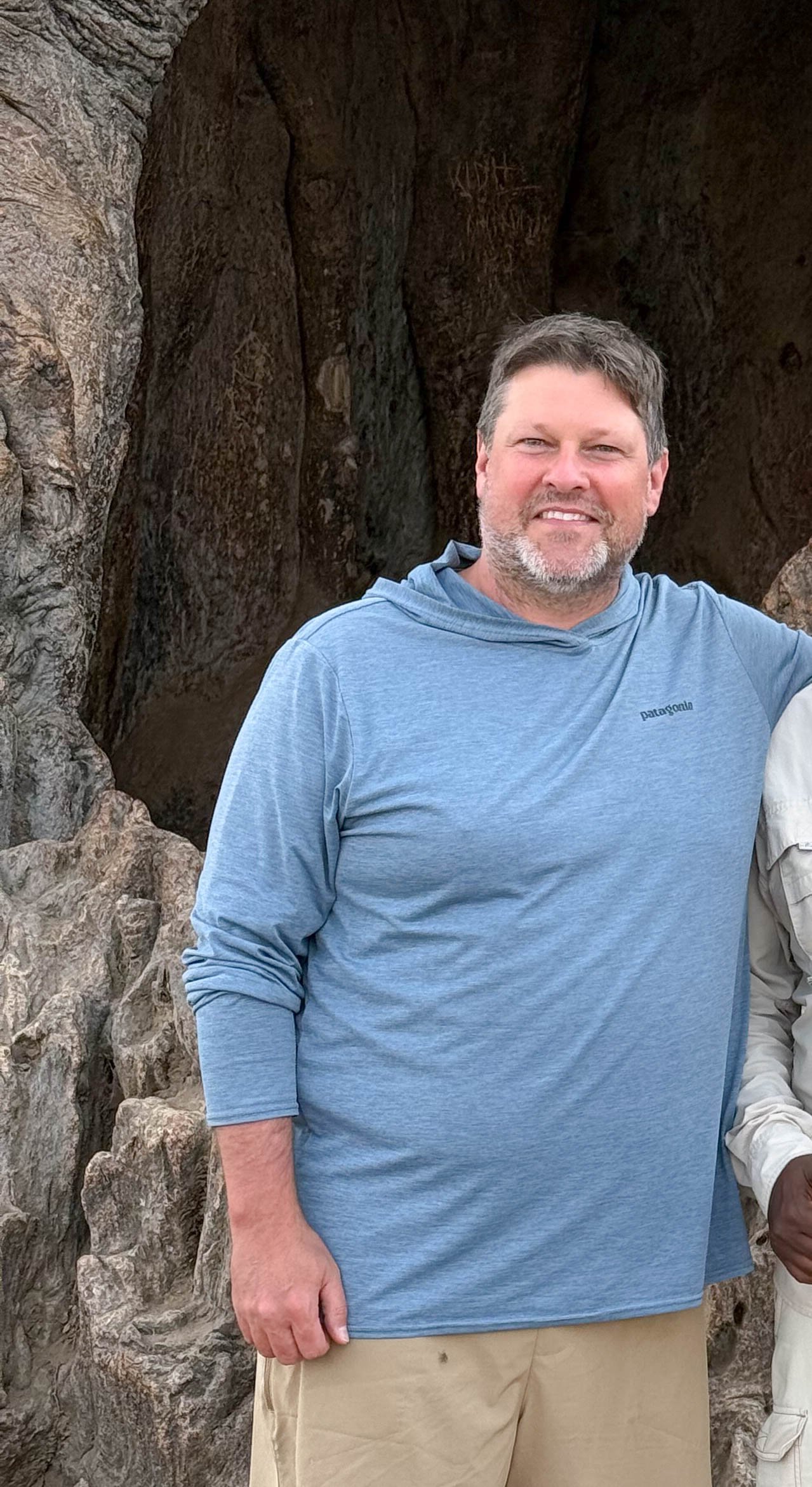 A man with a beard and mustache smiling, wearing a light blue Patagonia hoodie and beige pants, standing in front of a baobab tree.